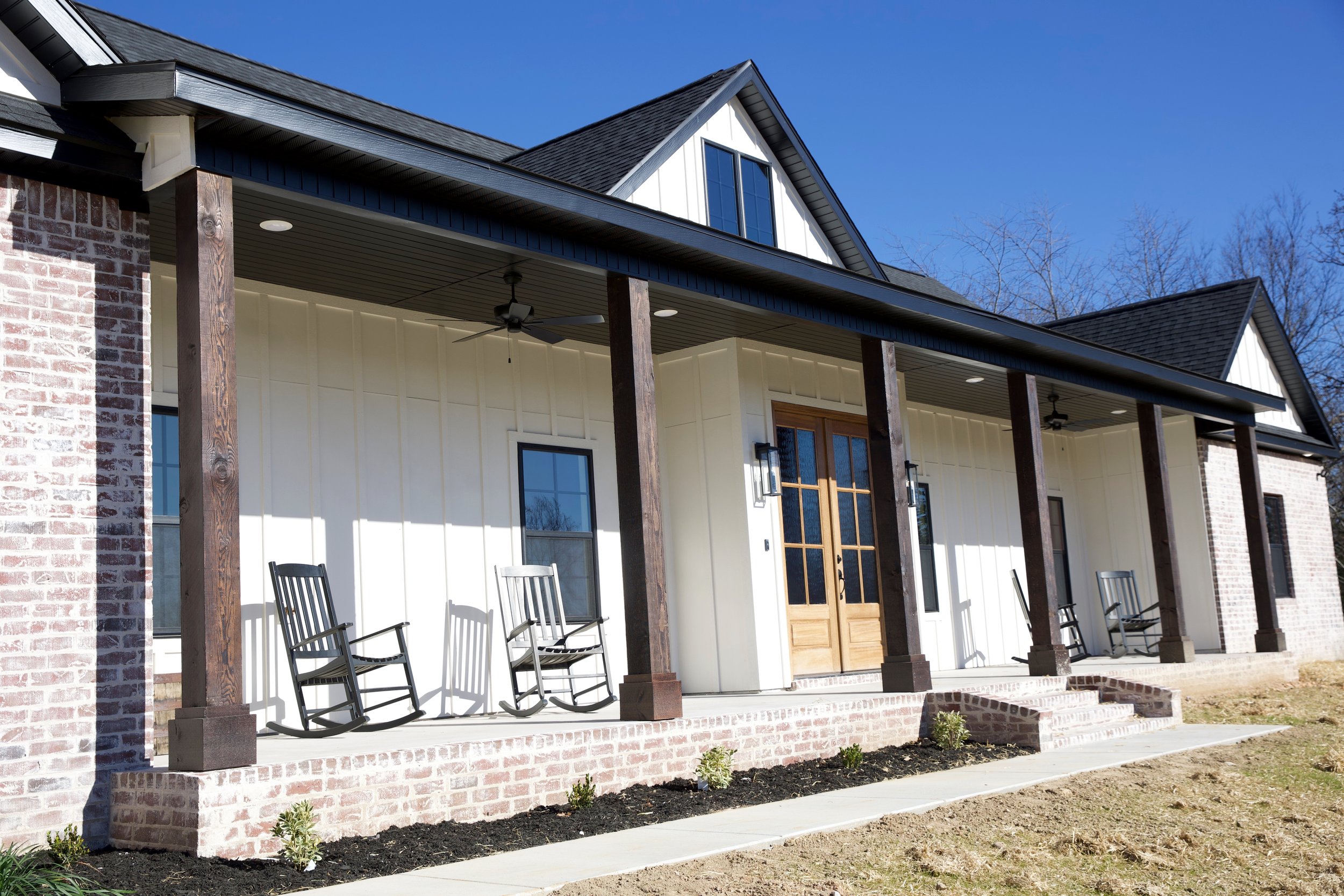 Front porch of a modern house with dark wood pillars, white siding, brick accents, and rocking chairs, under a gabled roof with dormer windows, in clear weather.