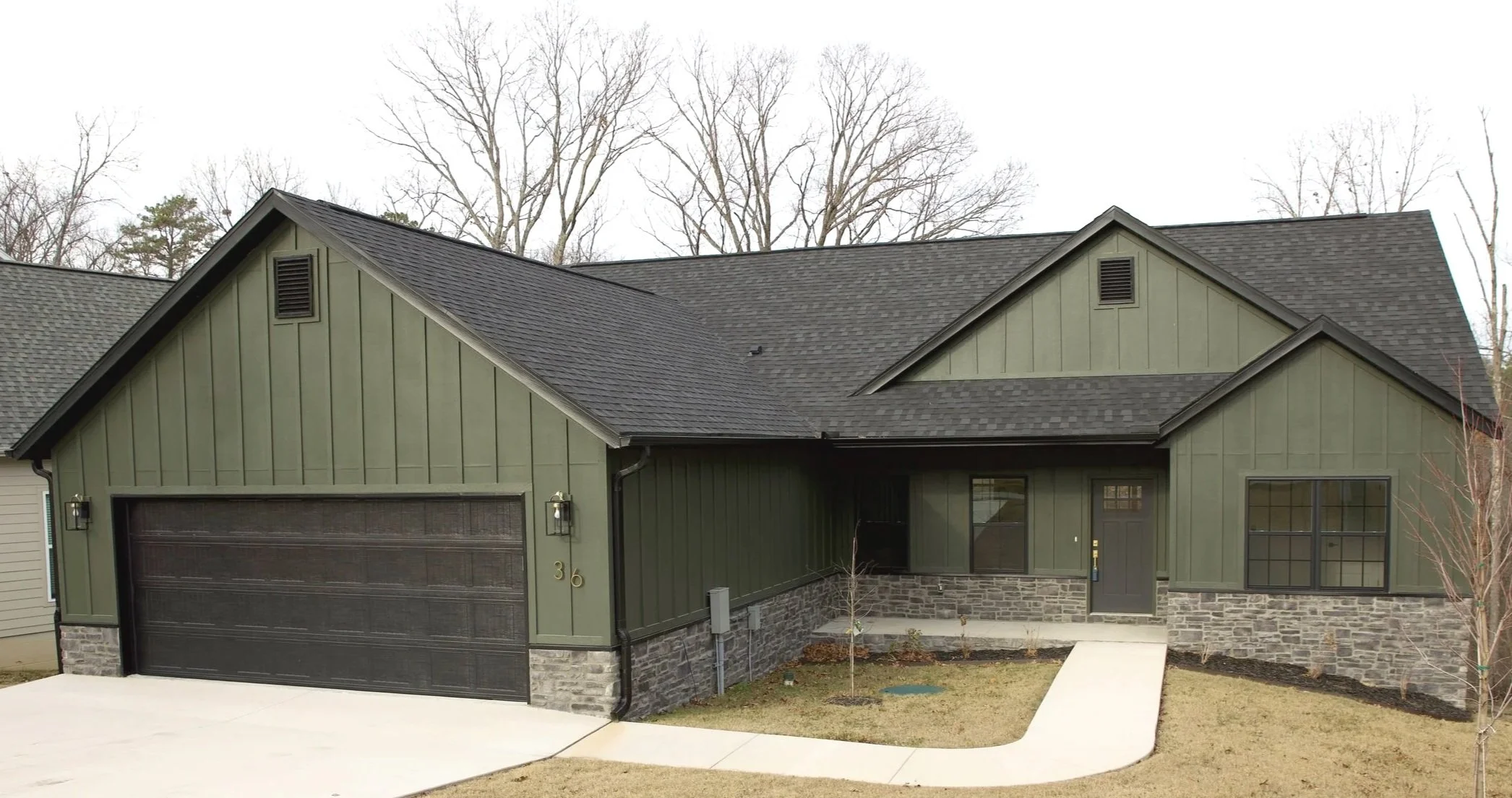 Modern house with green vertical siding, black garage door, stone accents, black window frames, and a concrete pathway leading to the front door, surrounded by leafless trees.