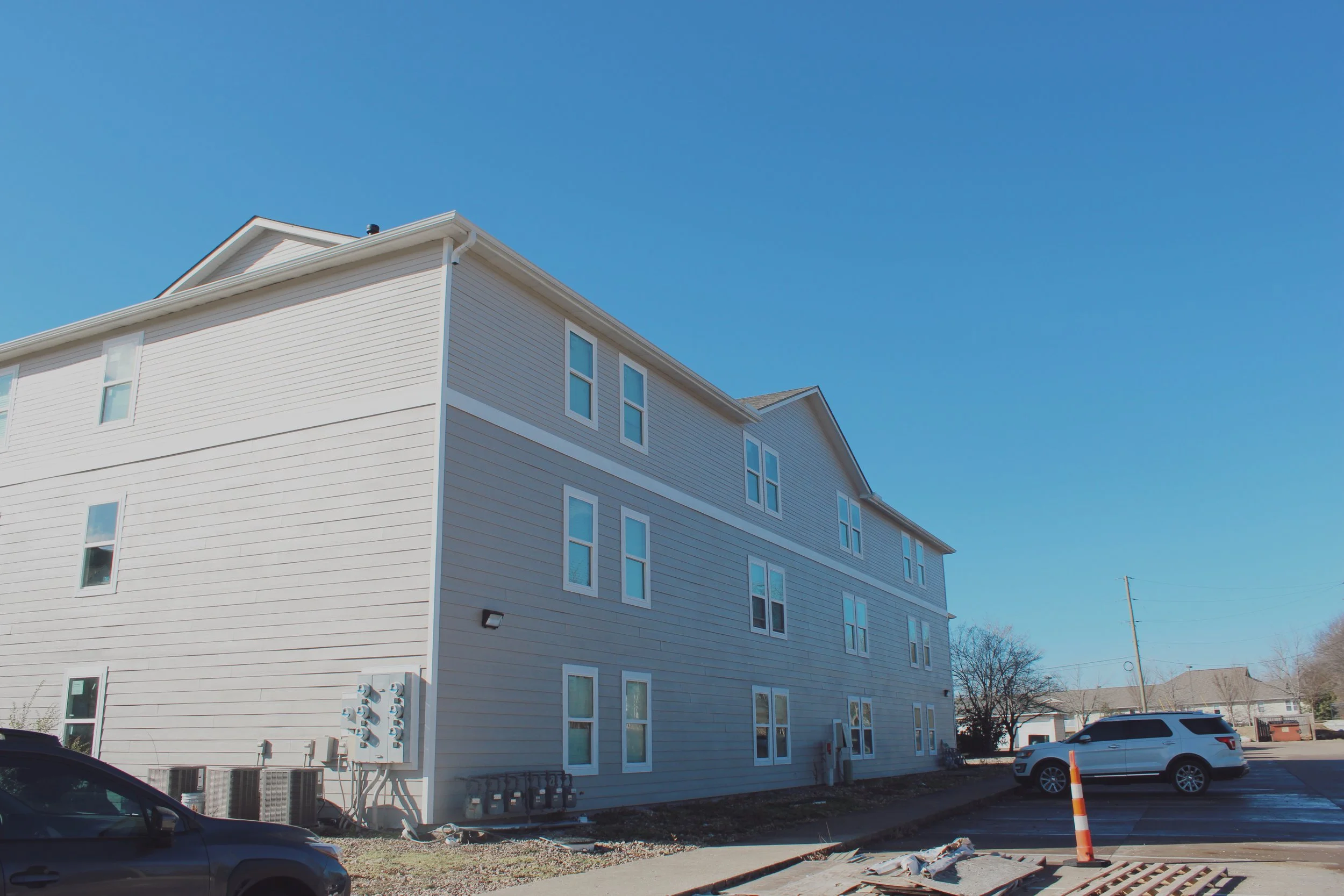 Exterior view of a multi-story white apartment building with multiple windows under a clear blue sky. There are parked cars and construction signs in the parking lot.