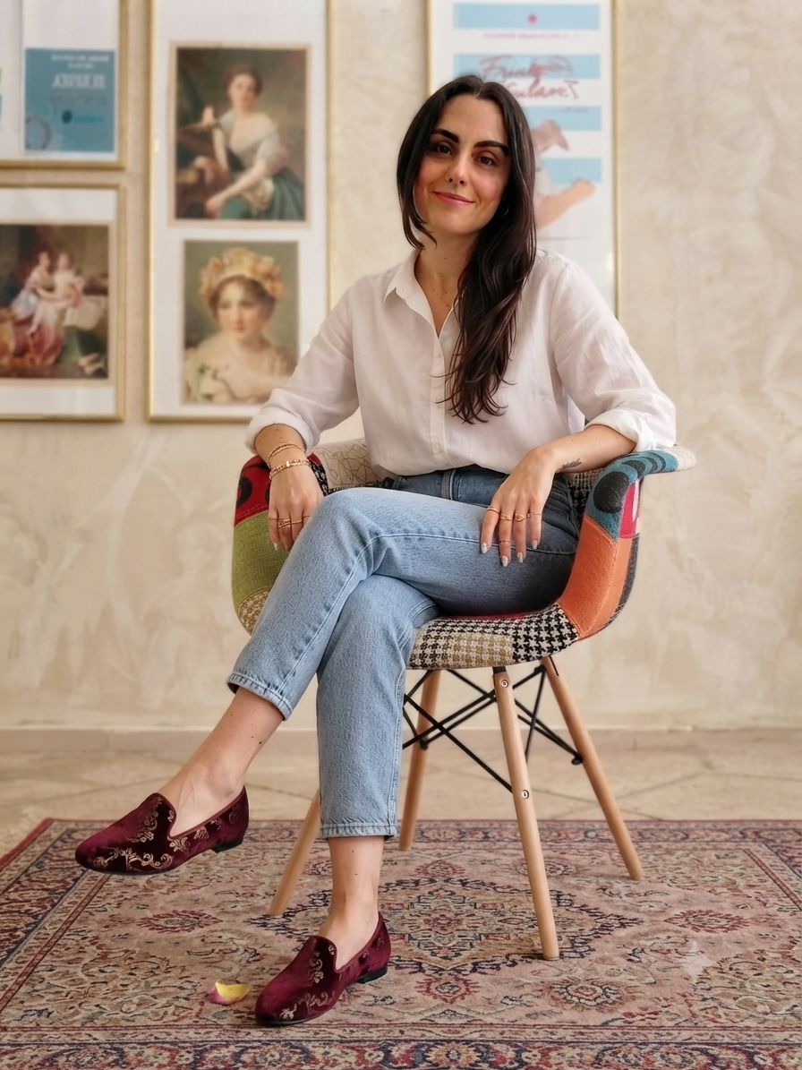 Donna seduta su una sedia colorata, indossa una camicia bianca e jeans azzurri, ha lunghi capelli castani e sorride, sullo sfondo ci sono quadri appesi alle pareti.