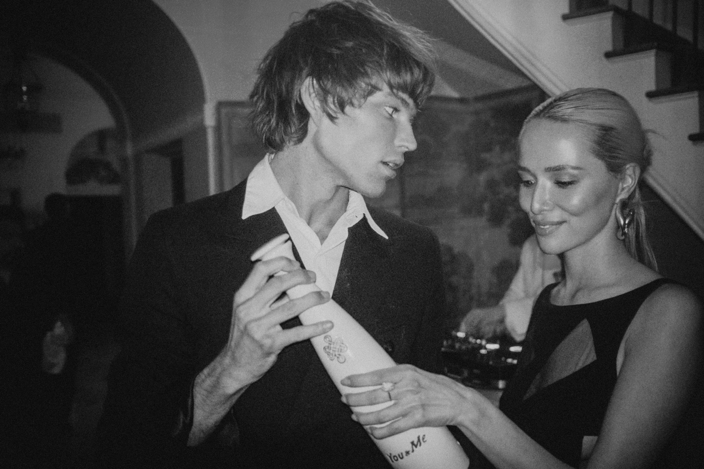 A man and woman stand close together at a social gathering, with the man holding a rolled-up piece of paper or possibly a bottle. Both are dressed in semi-formal attire, the man in a suit and white shirt, and the woman in a sleeveless black dress with earrings. They are smiling softly at each other, with the woman placing her hand on the man's chest or sleeve. The background shows an indoor setting with a staircase and a person in the background.