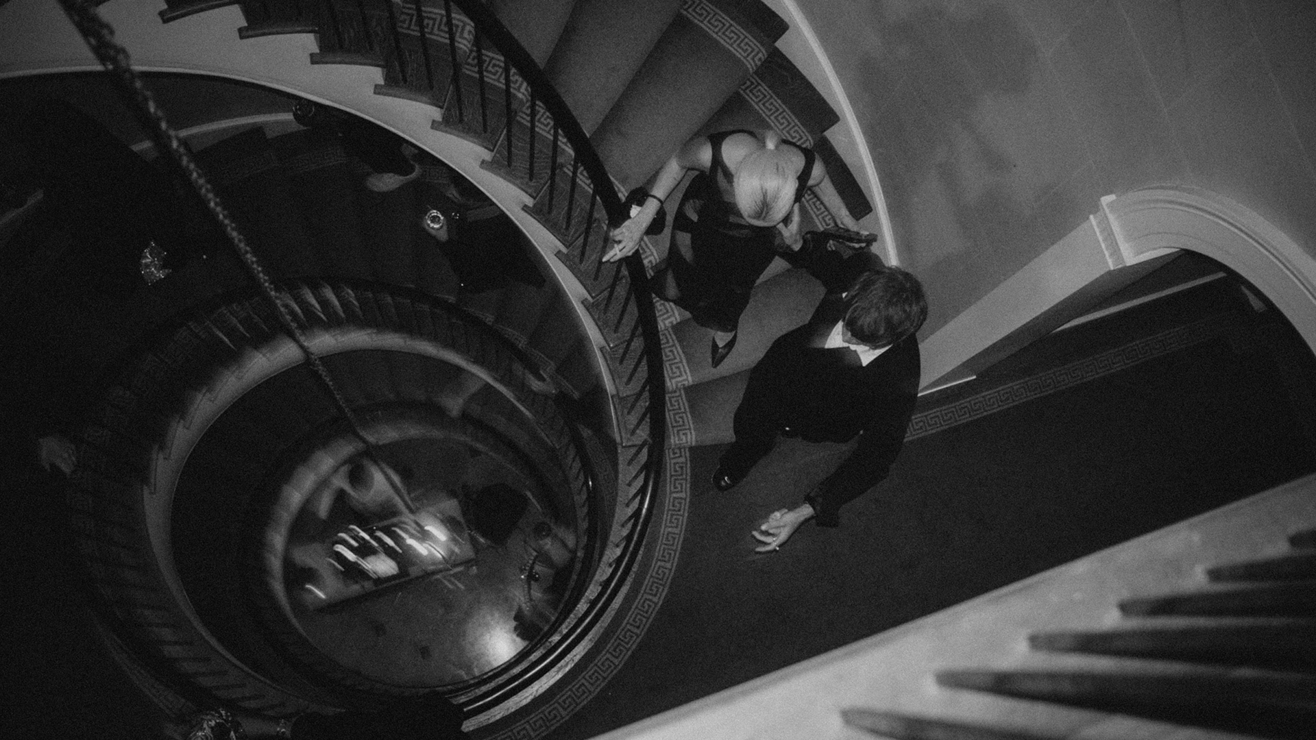 A black and white top-down photo of a woman in a dress and a man in a suit walking down a spiral staircase with ornate details.