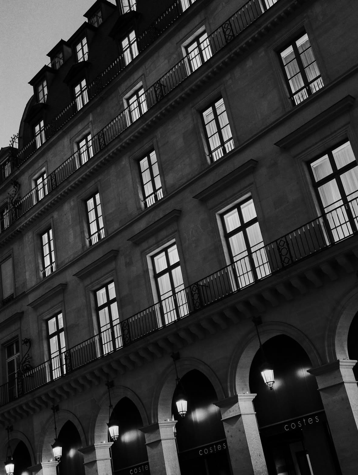 Black and white photo of a multi-story building with arched entrances on the ground level, lit lamps hanging from the arches, and multiple windows with balconies above.