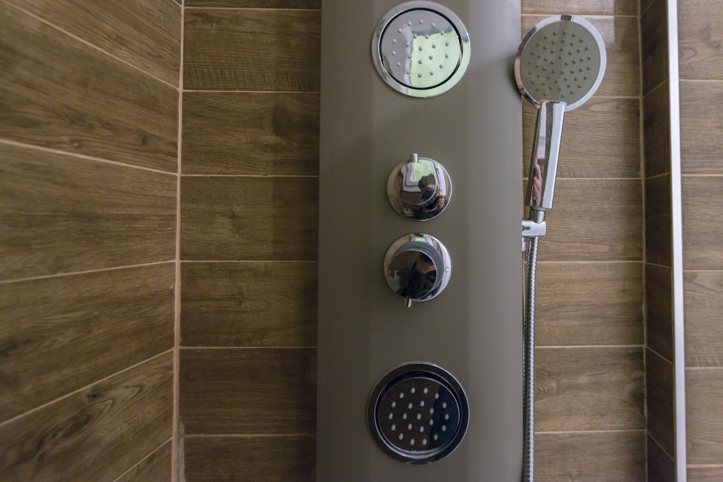 Vertical view of a shower control panel with three round chrome knobs and two round showerheads, installed on a wall with brown wooden tiles.