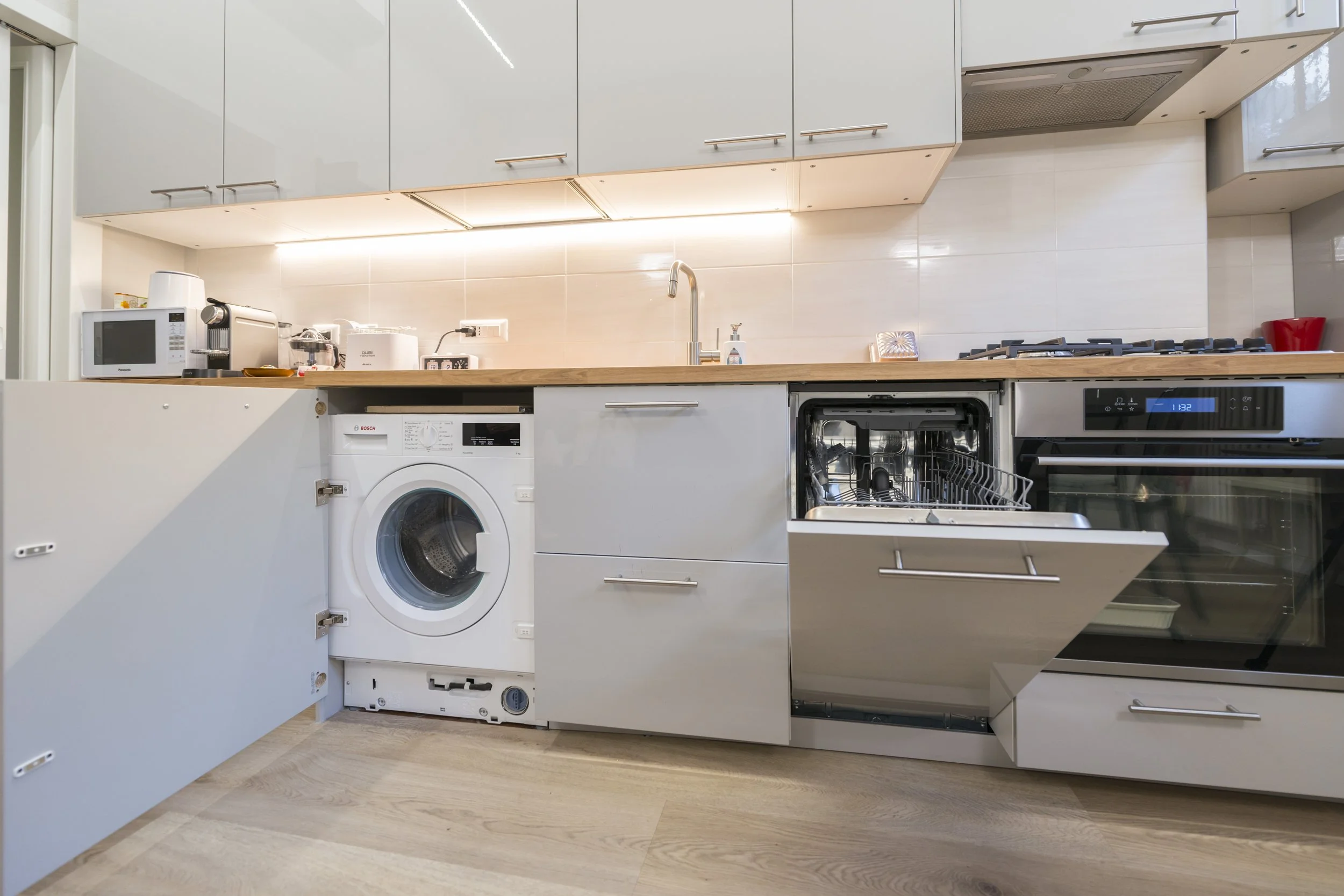 Modern kitchen with open dishwasher, washing machine, microwave, oven, and various small appliances on the countertop.