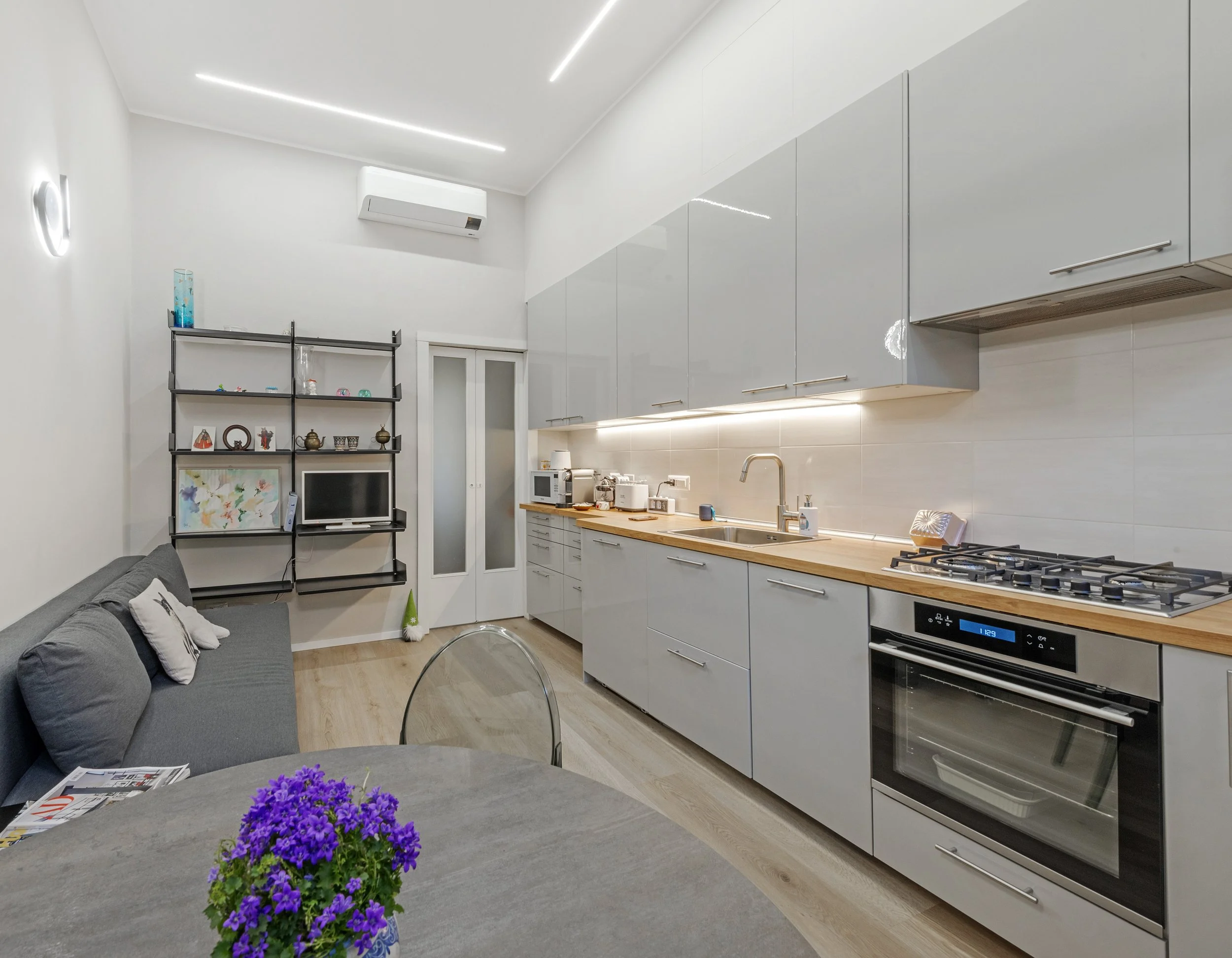 Modern kitchen with white cabinets, wooden countertop, gas stove, oven, sink, microwave, and small living area with a gray sofa and a black shelf with decorative items.