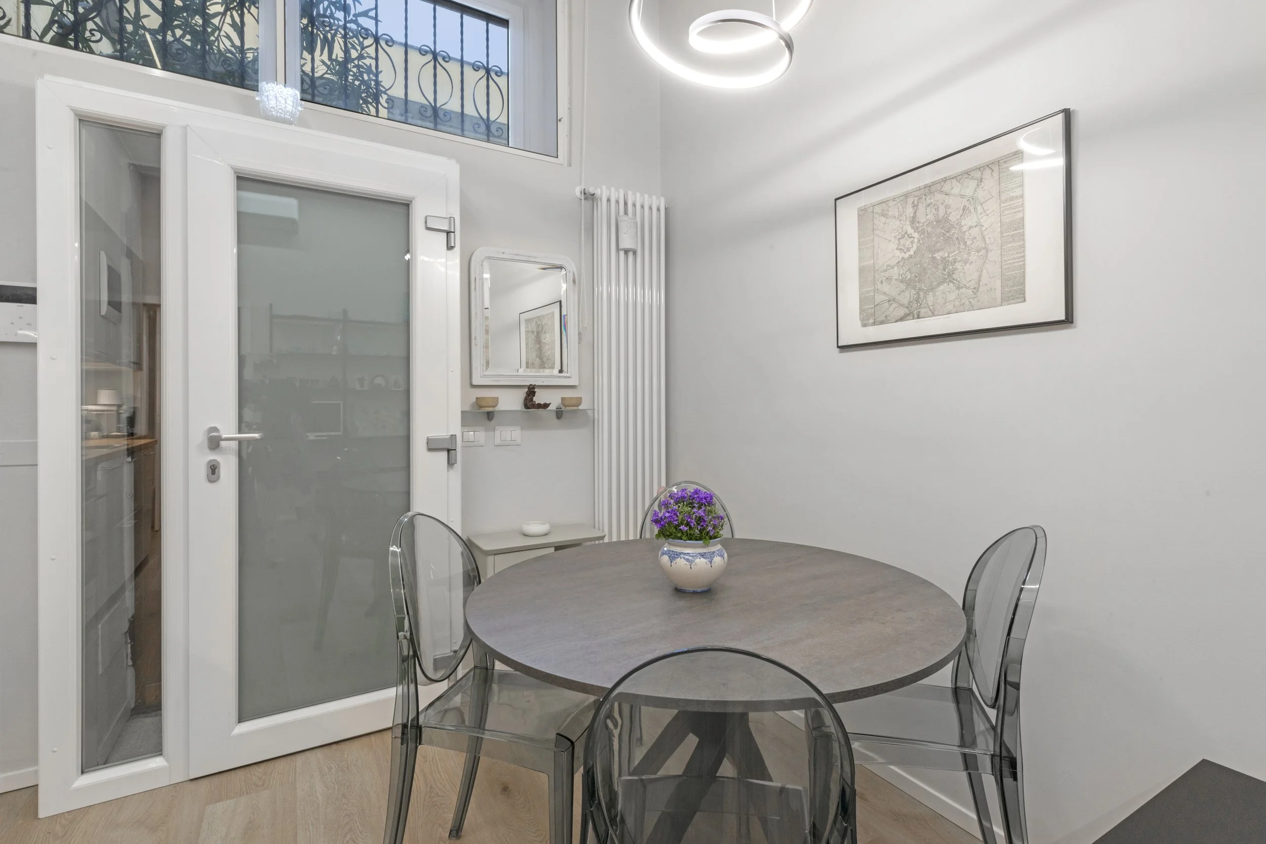 A modern dining room featuring a round wooden table, four transparent chairs, and a potted purple flower centerpiece. The room has white walls, a framed map, a white radiator, a mirror above a small shelf, and a circular ceiling light.