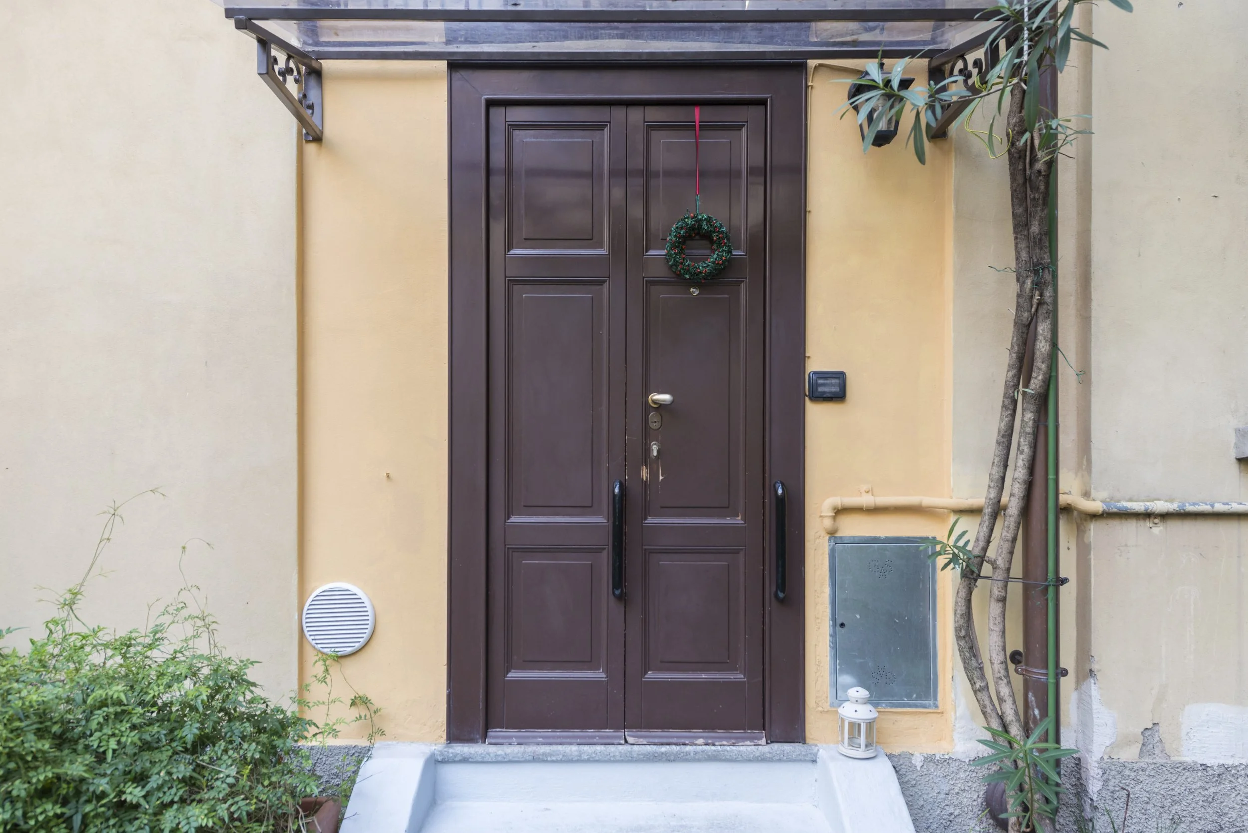 Brown front door with a Christmas wreath hanging on it, set within a yellow wall, with a small lantern on the ground and a plant growing on the right side.