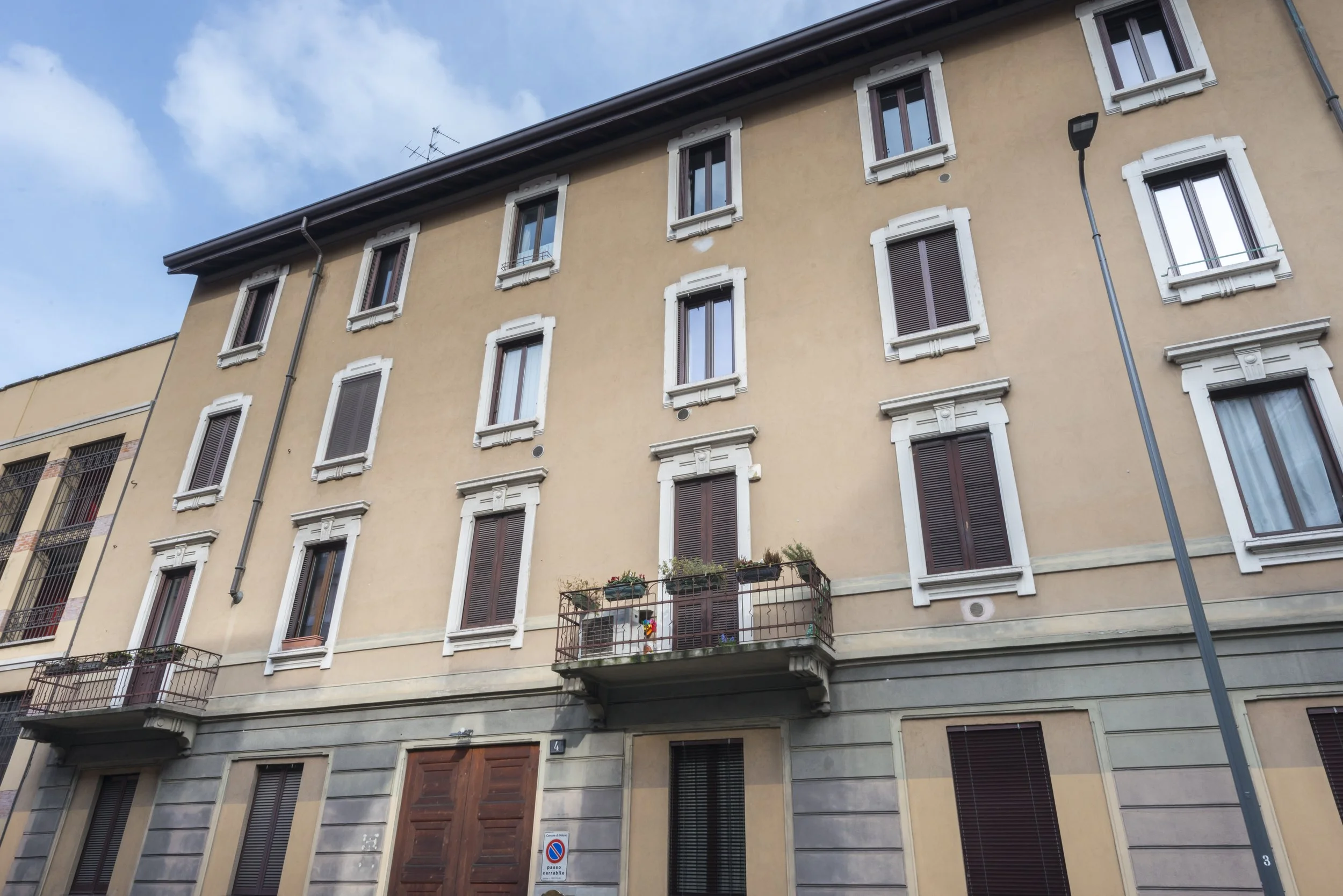 Four-story beige building with brown shutters and small balconies with potted plants. Street lamp in foreground and blue sky with clouds.
