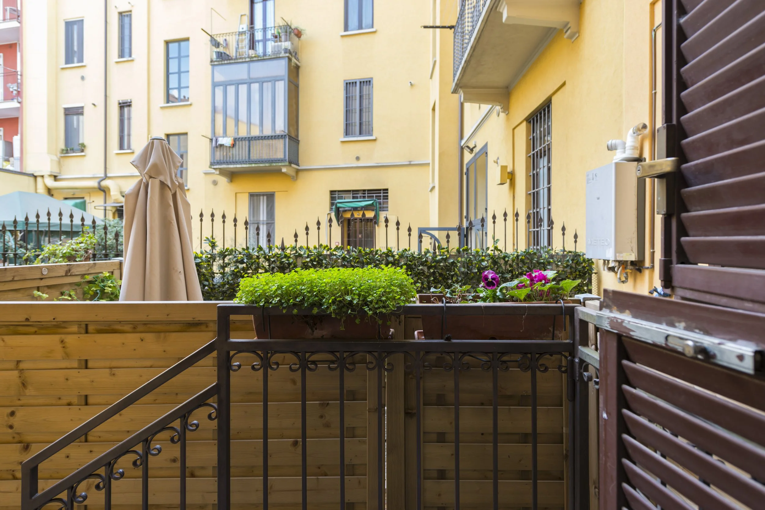 View of a small apartment balcony with flower pots and a closed beige umbrella, overlooking neighboring yellow buildings with windows, balconies, and outdoor furniture.