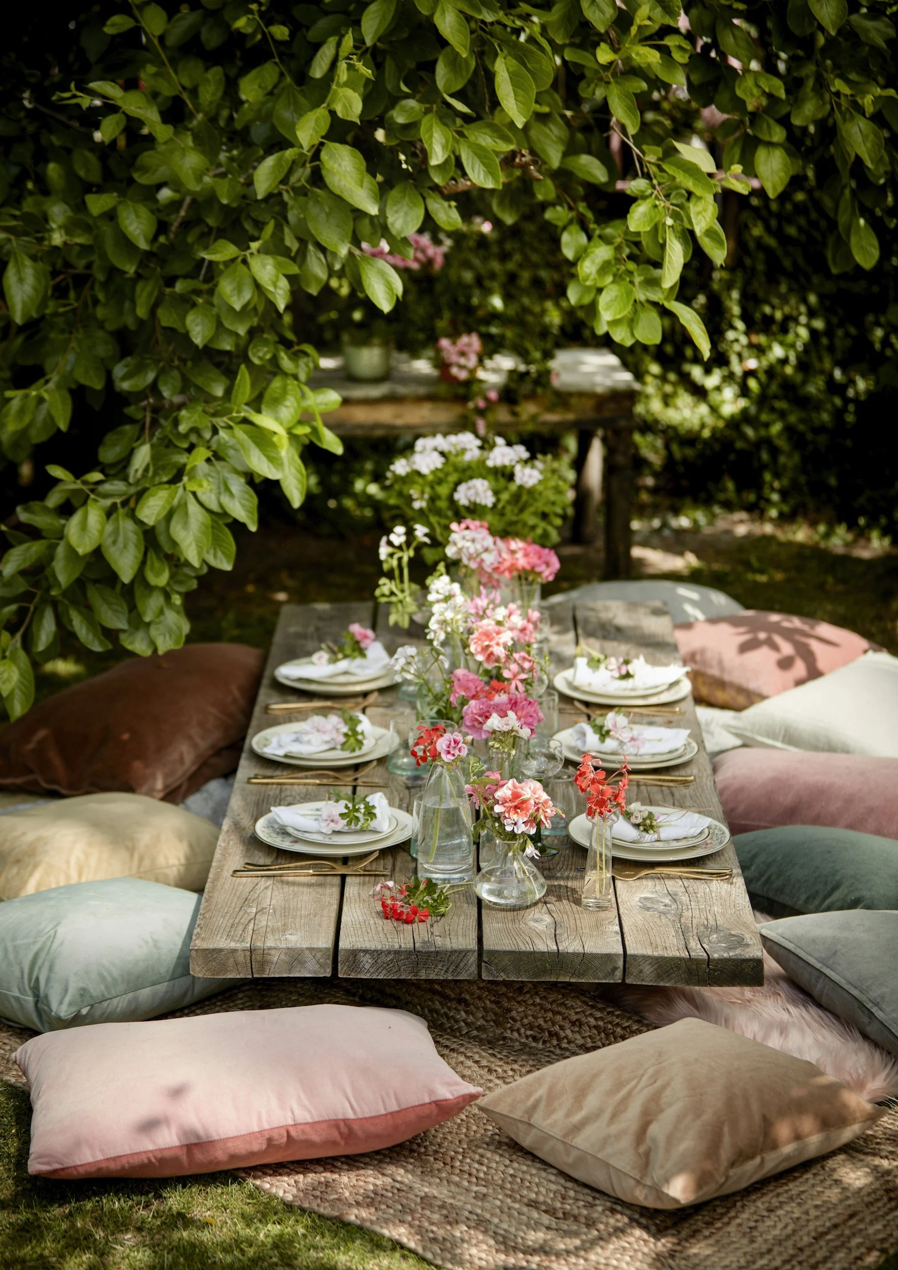 A rustic outdoor dining setup on a wooden table with floral arrangements in glass vases, surrounded by pastel-colored pillows and cushions on a woven rug among lush green foliage.