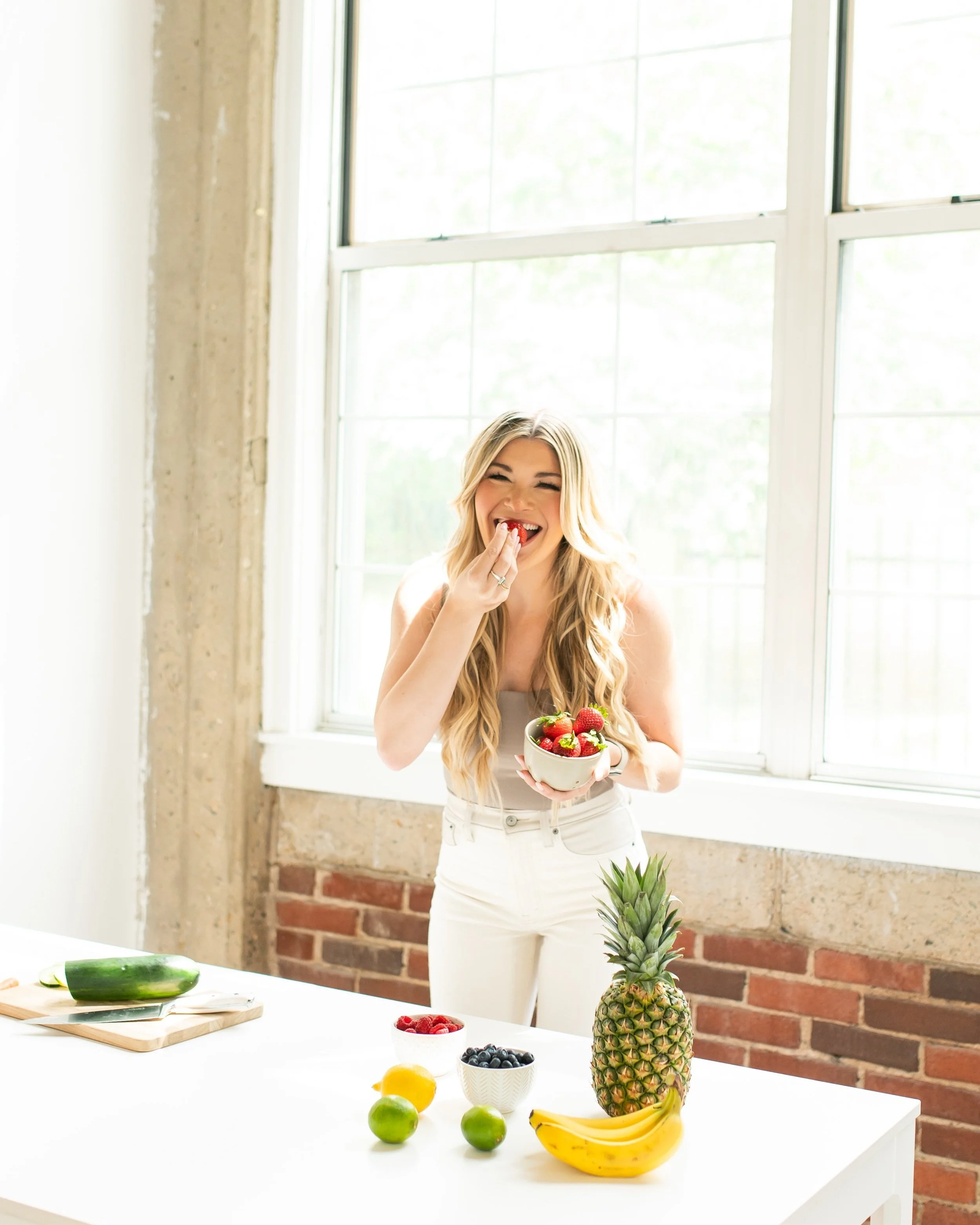 Haley eating a strawberry bowl in a bright kitchen with large windows, surrounded by various fruits including pineapple, bananas, blueberries, raspberries, and limes.