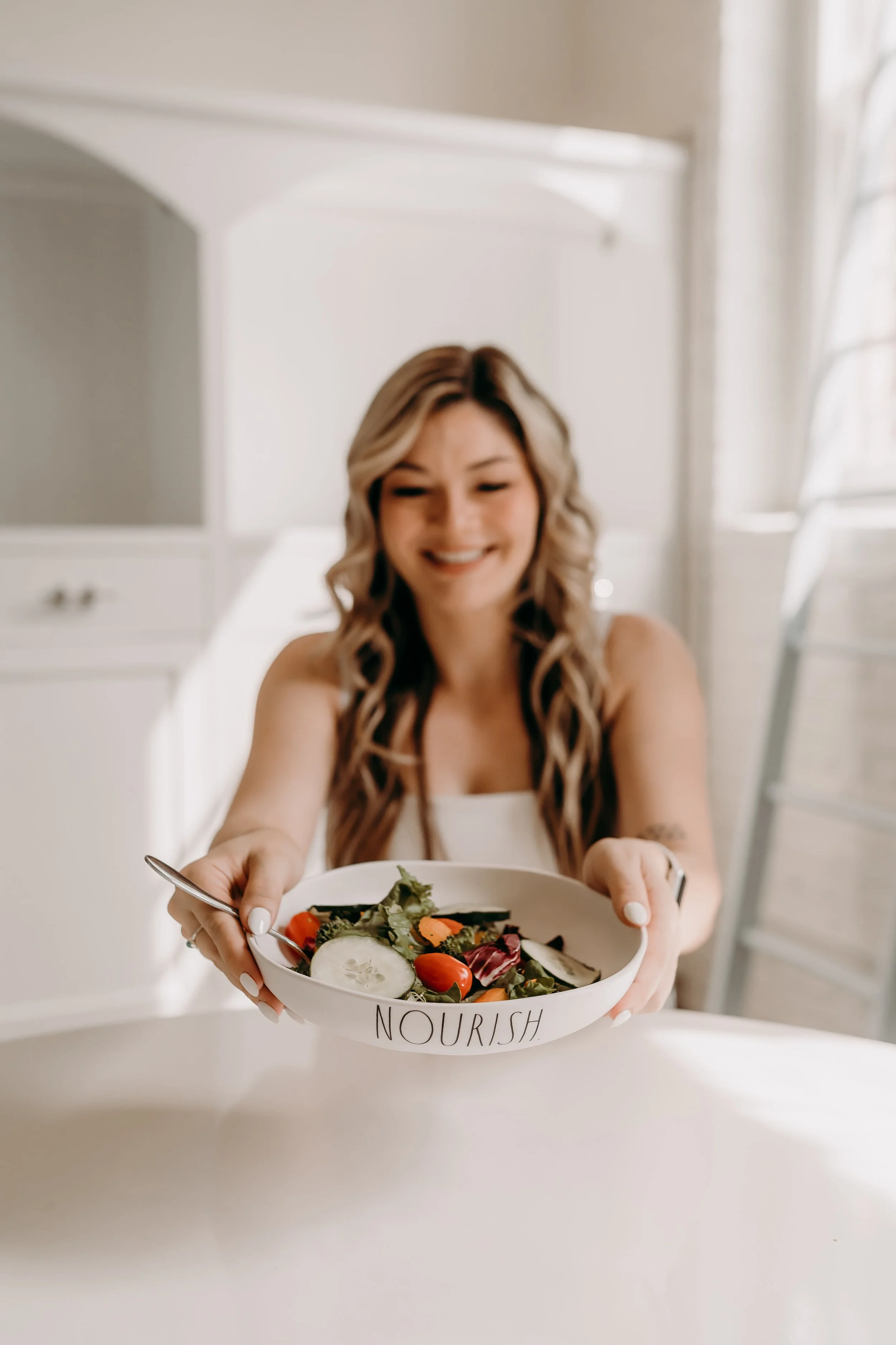 Smiling woman holding a bowl of salad with the word "NOURISH" written on it, indoors near a window.