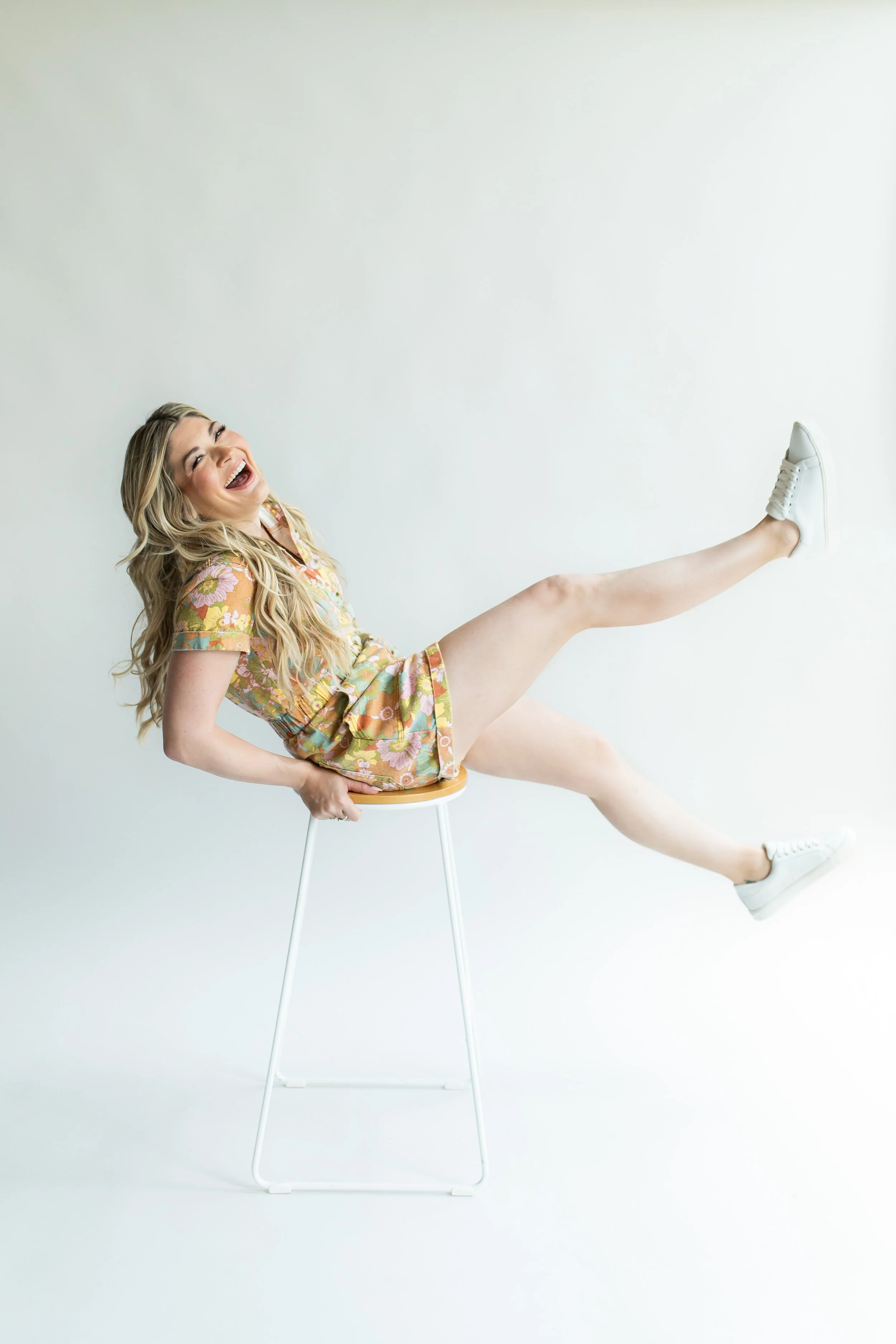 A woman with long blonde hair laughing while sitting sideways on a chair with her back against the wall, wearing a floral dress and white sneakers with one leg extended upward.