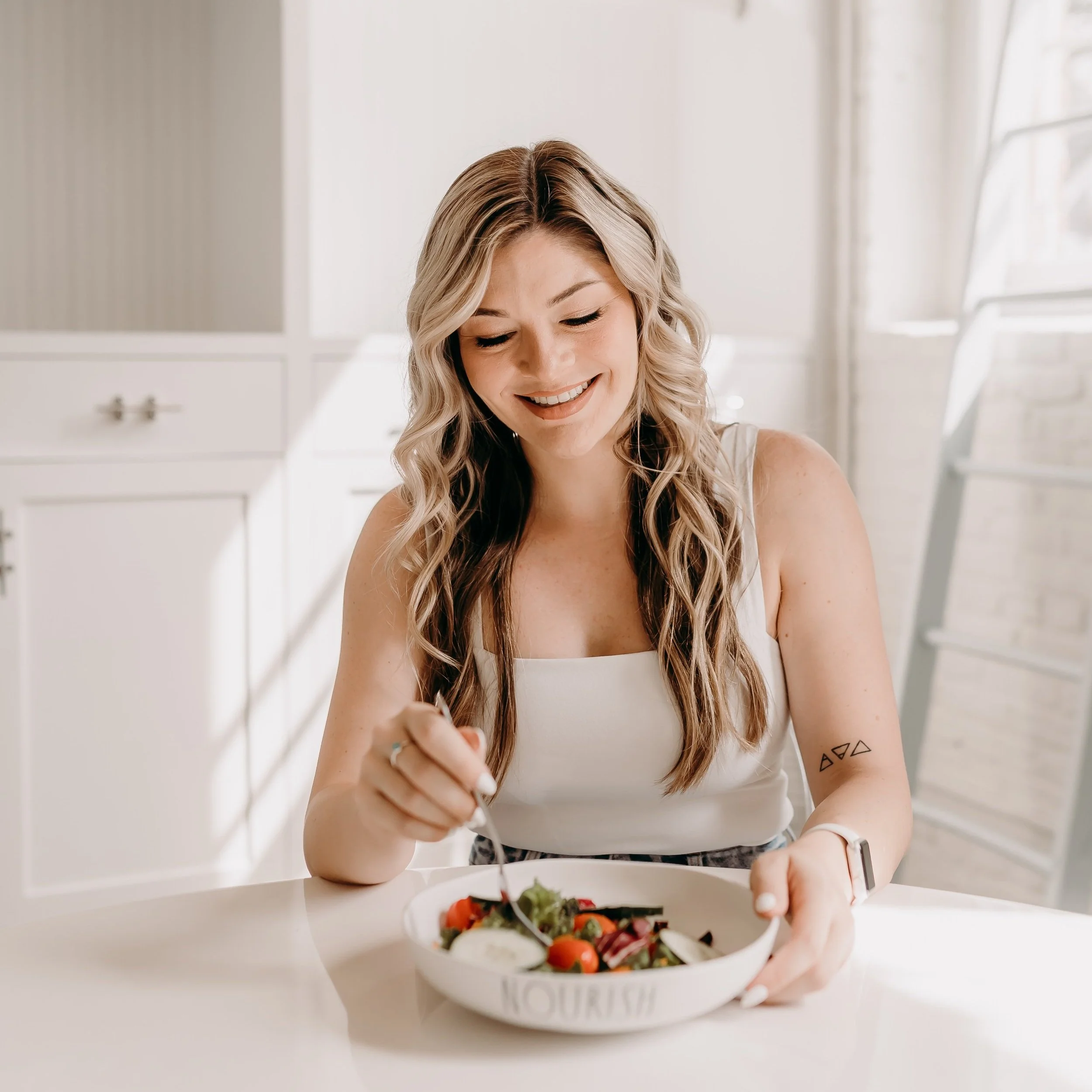 A woman with long wavy blonde hair sitting at a table, smiling while eating a fresh salad in a bright, modern kitchen.