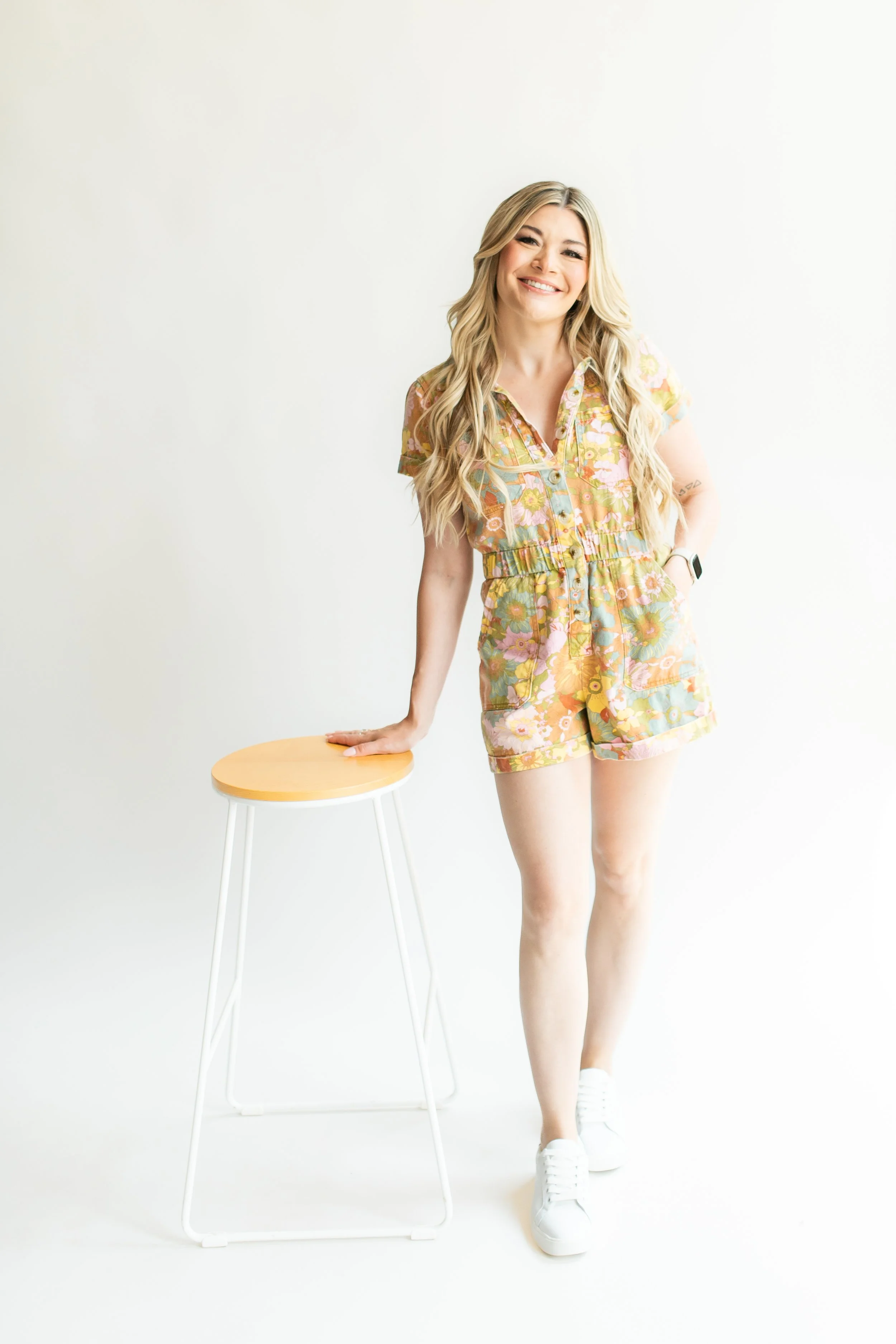 A young woman with long blonde hair in a floral romper, smiling and standing next to a white stool with a light wood top, against a plain white background.