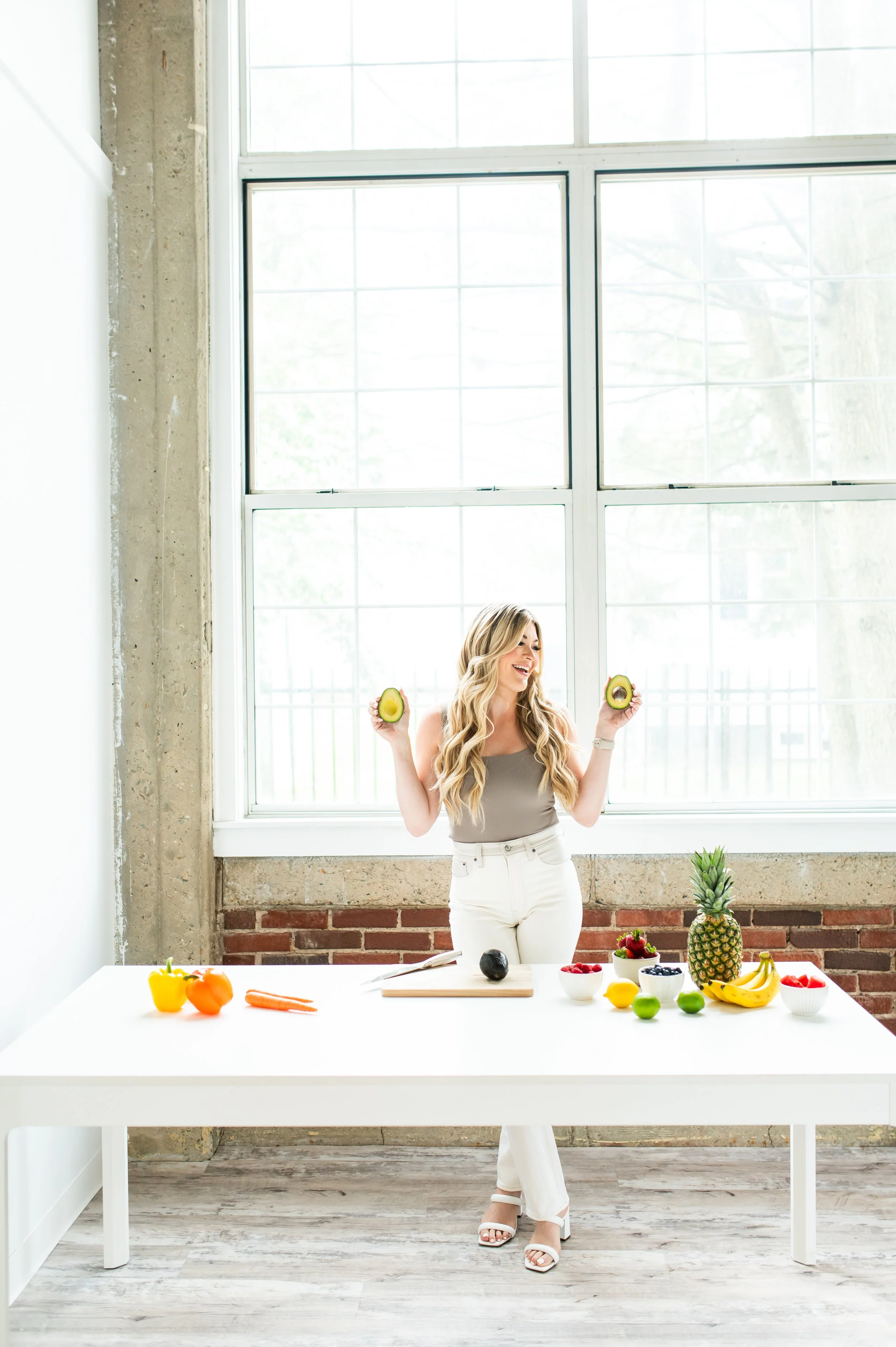 A woman with long blonde hair and white pants holding cut avocados in front of a large window. A white table in front of her displays various fruits and vegetables including a pineapple, bananas, strawberries, blueberries, a lemon, a green apple, a bowl of raspberries, and bell peppers.