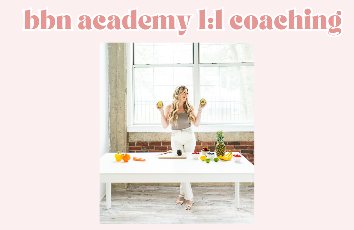 Woman in gray tank top and white pants holding avocados, standing behind a white table with fruits and vegetables, in a bright room with a large window and brick accent wall, promoting BHN Academy's 1:1 coaching.