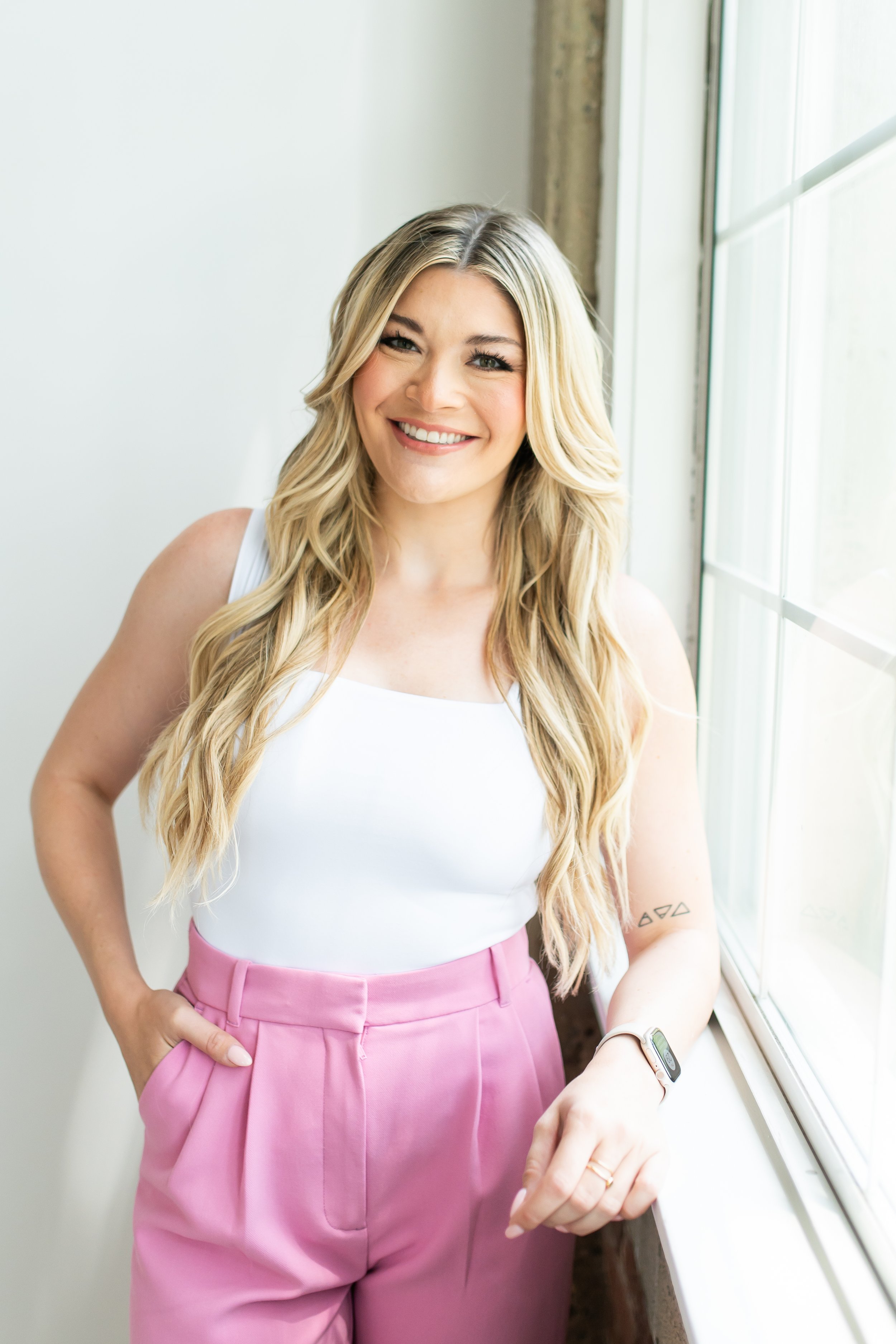 A young woman with long blonde hair smiling and looking at the camera, standing near a window with natural light.