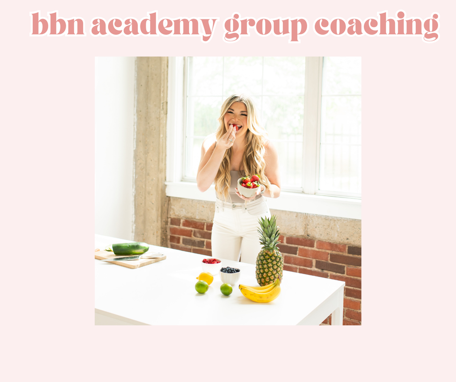 A woman with long blonde hair eating strawberries from a bowl in a bright kitchen. There are fresh fruits on the white counter, including a pineapple, bananas, lemons, limes, blueberries, and strawberries.
