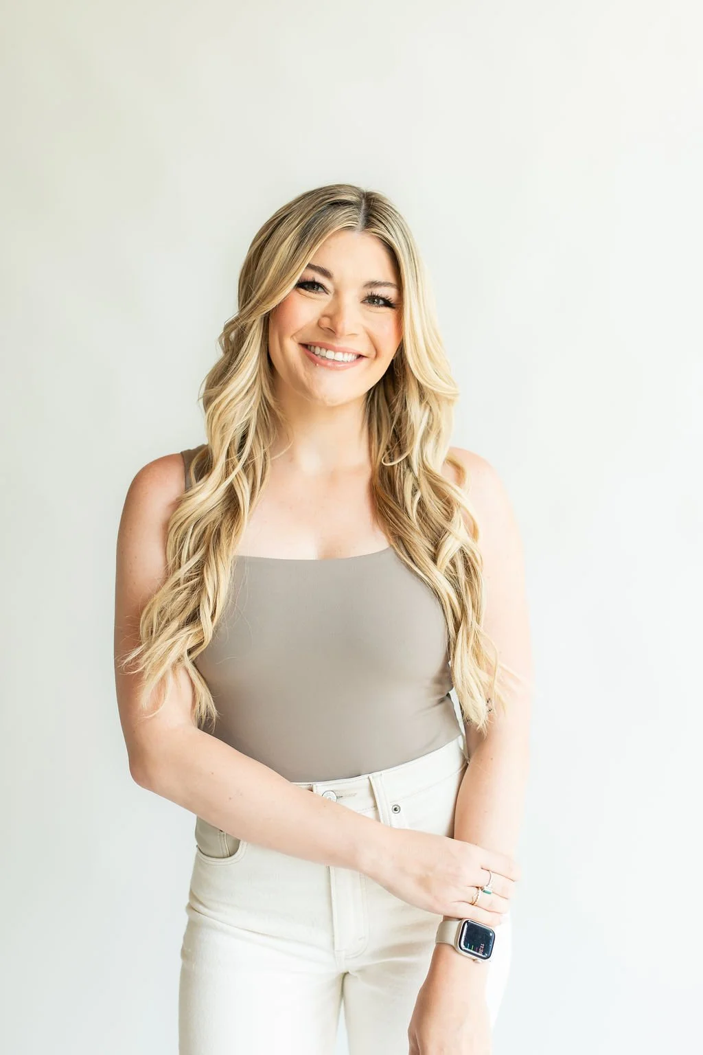 A smiling woman with long blonde wavy hair wearing a beige sleeveless top and white pants, standing against a plain light background, with an Apple Watch on her left wrist.