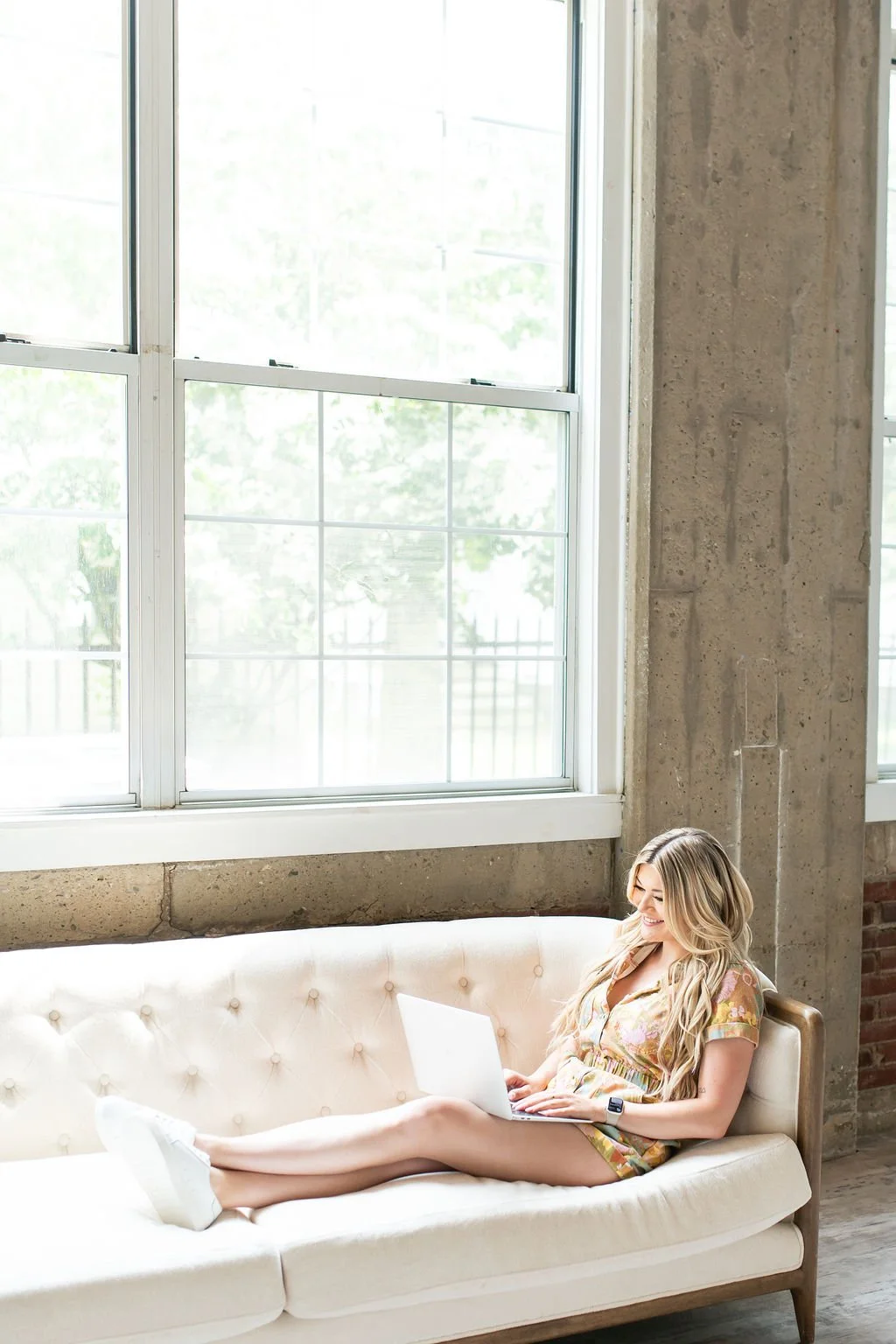 Haley sitting on a cream-colored tufted sofa, using a laptop, next to large windows with a cityscape view.