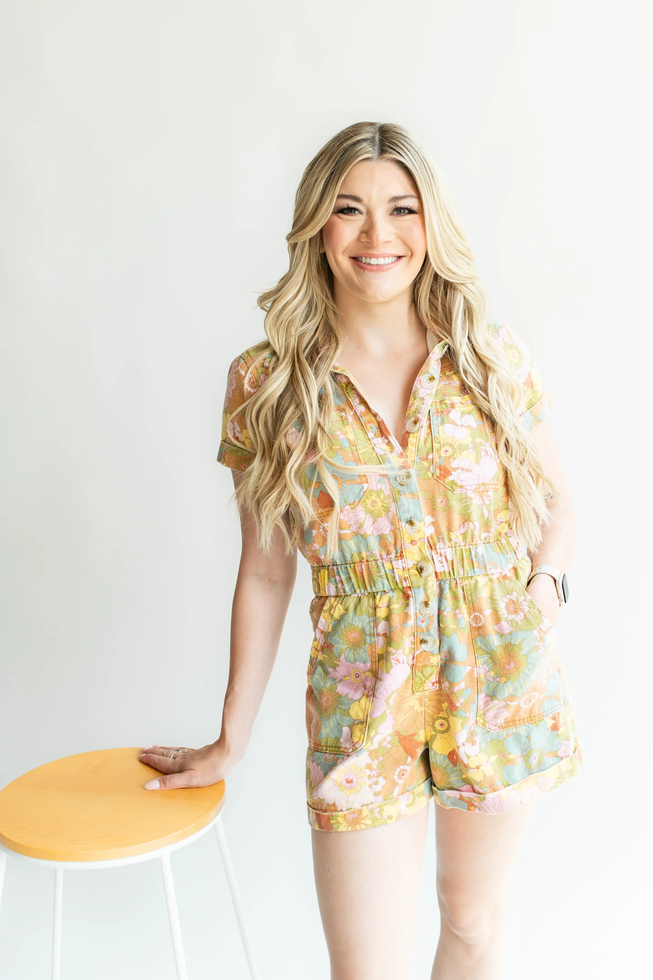 A smiling woman with long blond hair wearing a colorful floral romper, standing with one hand on a wooden stool against a plain white background.