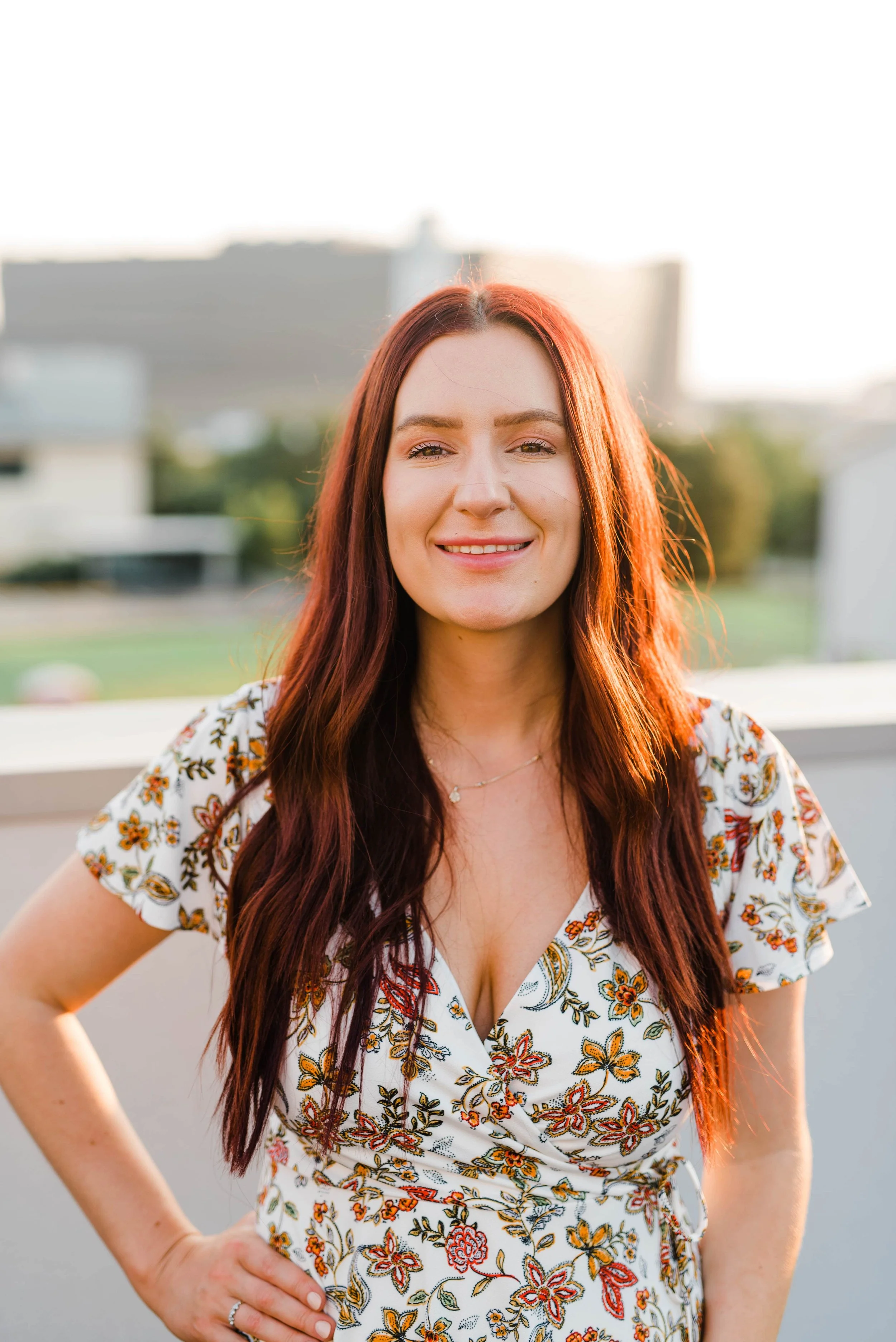 A young woman with long red hair smiling outdoors at sunset, wearing a floral dress.
