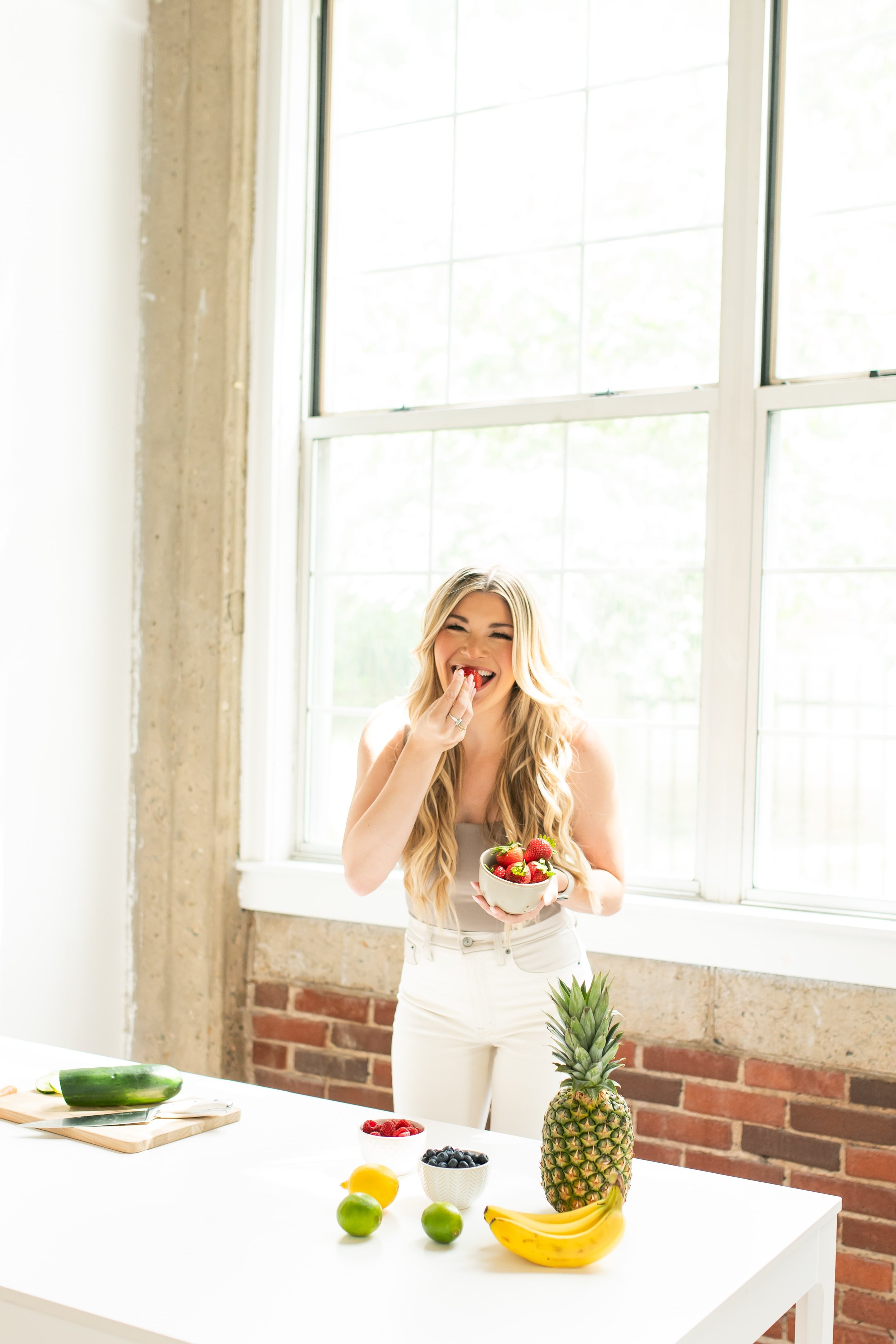 Haley laughing and holding a bowl of strawberries in a bright kitchen with a white table and various fruits including pineapple, bananas, blueberries, strawberries, a lemon, and limes.