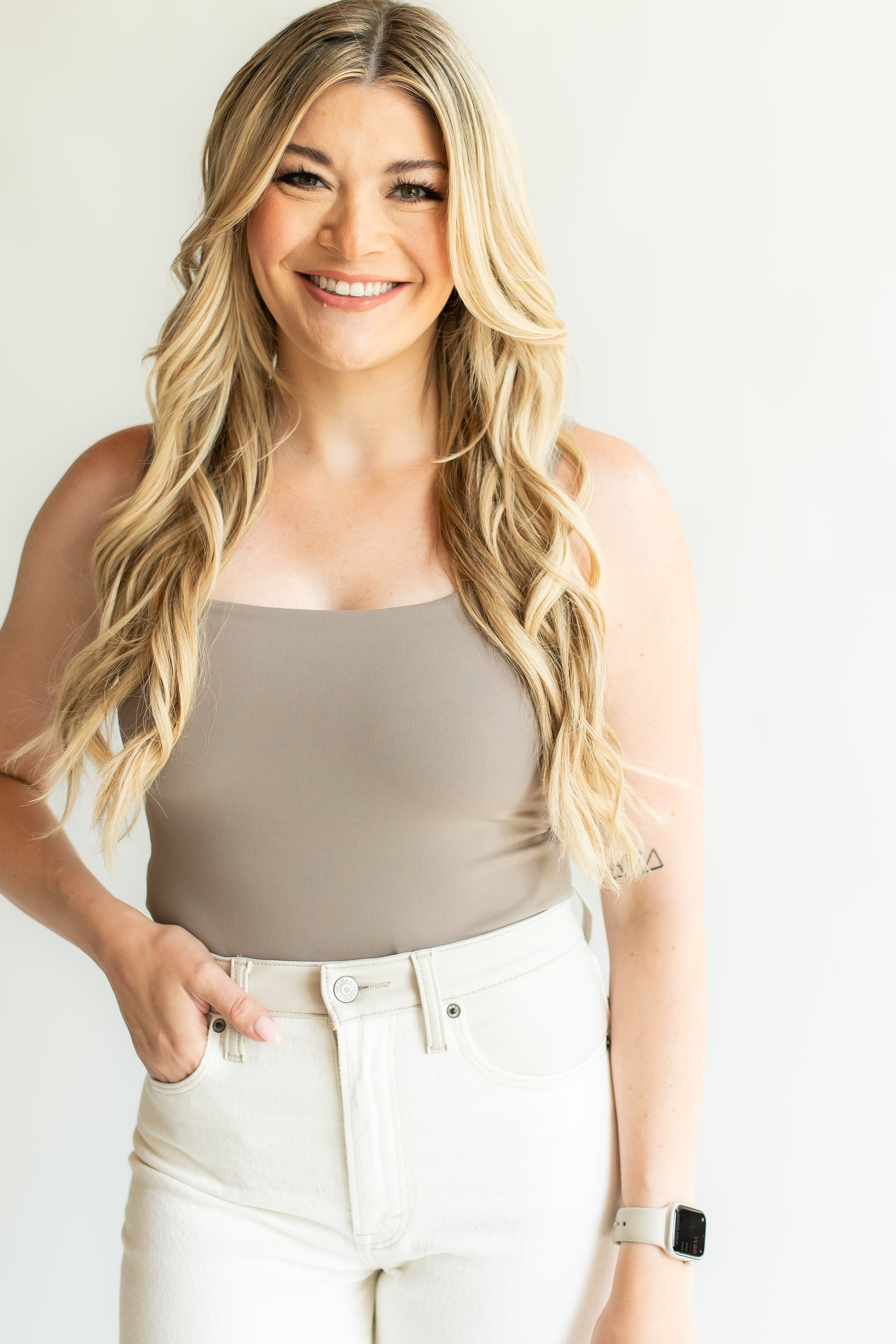 Haley wearing a beige sleeveless top, white pants, and a smartwatch, standing against a plain light background.