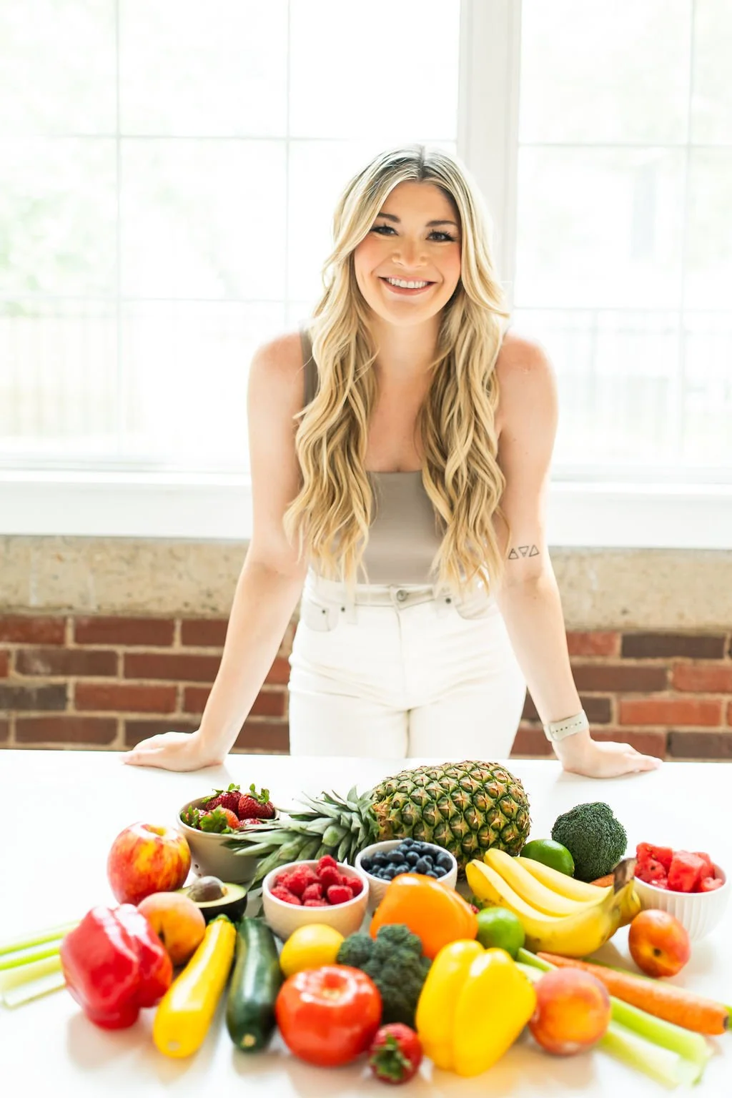 Young woman with long blonde hair smiling at the camera, standing behind a table filled with various fresh fruits and vegetables in front of a bright window.