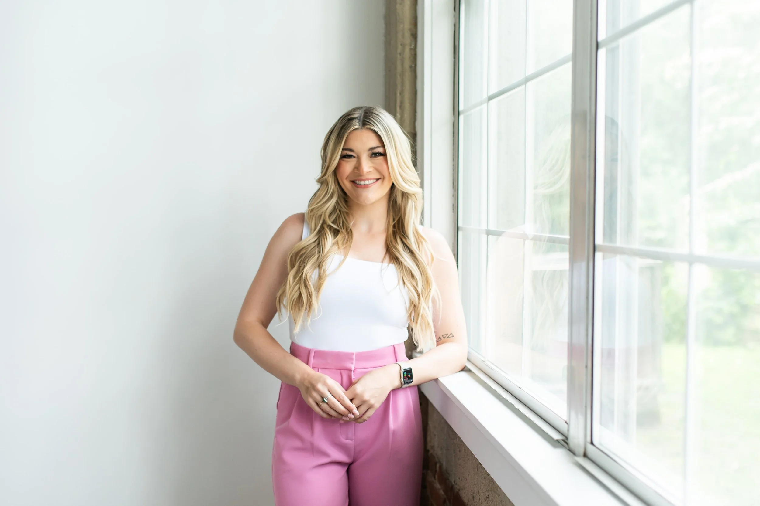 A young woman with long blonde hair, wearing a white sleeveless top and pink high-waisted pants, standing near a large window with greenery outside, smiling and looking at the camera.