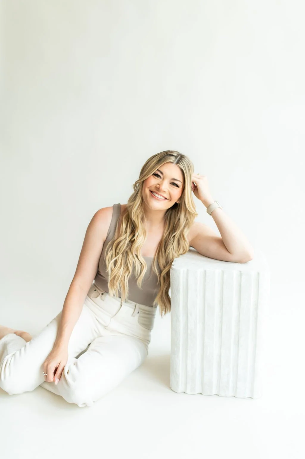 A smiling woman with long blonde hair, wearing a taupe sleeveless top and white pants, sitting on the floor with her left arm resting on a white marble pedestal and her right hand on her knee, against a plain white background.
