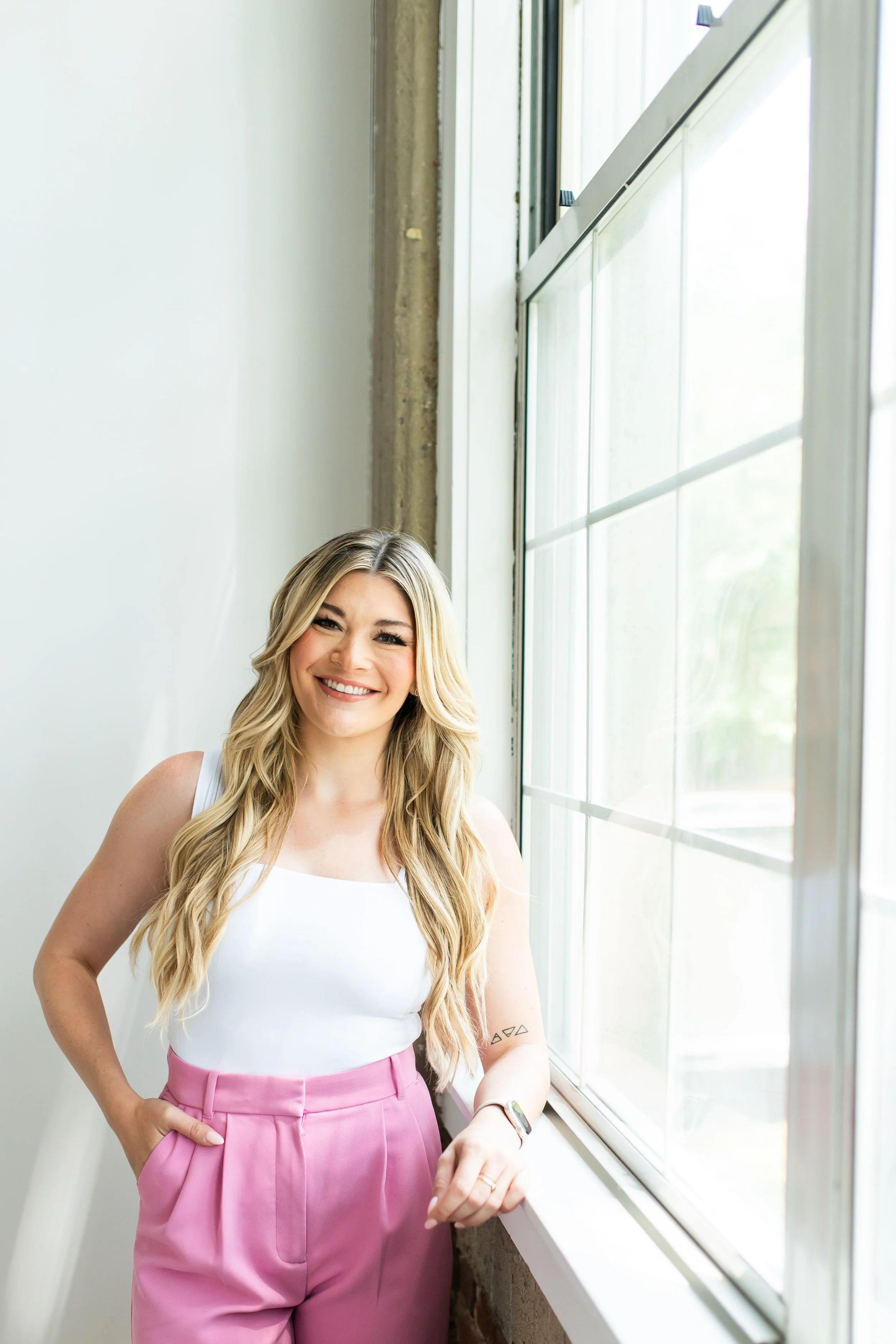 Haley standing next to a large window in a bright room, wearing a white sleeveless top and pink high-waisted pants.