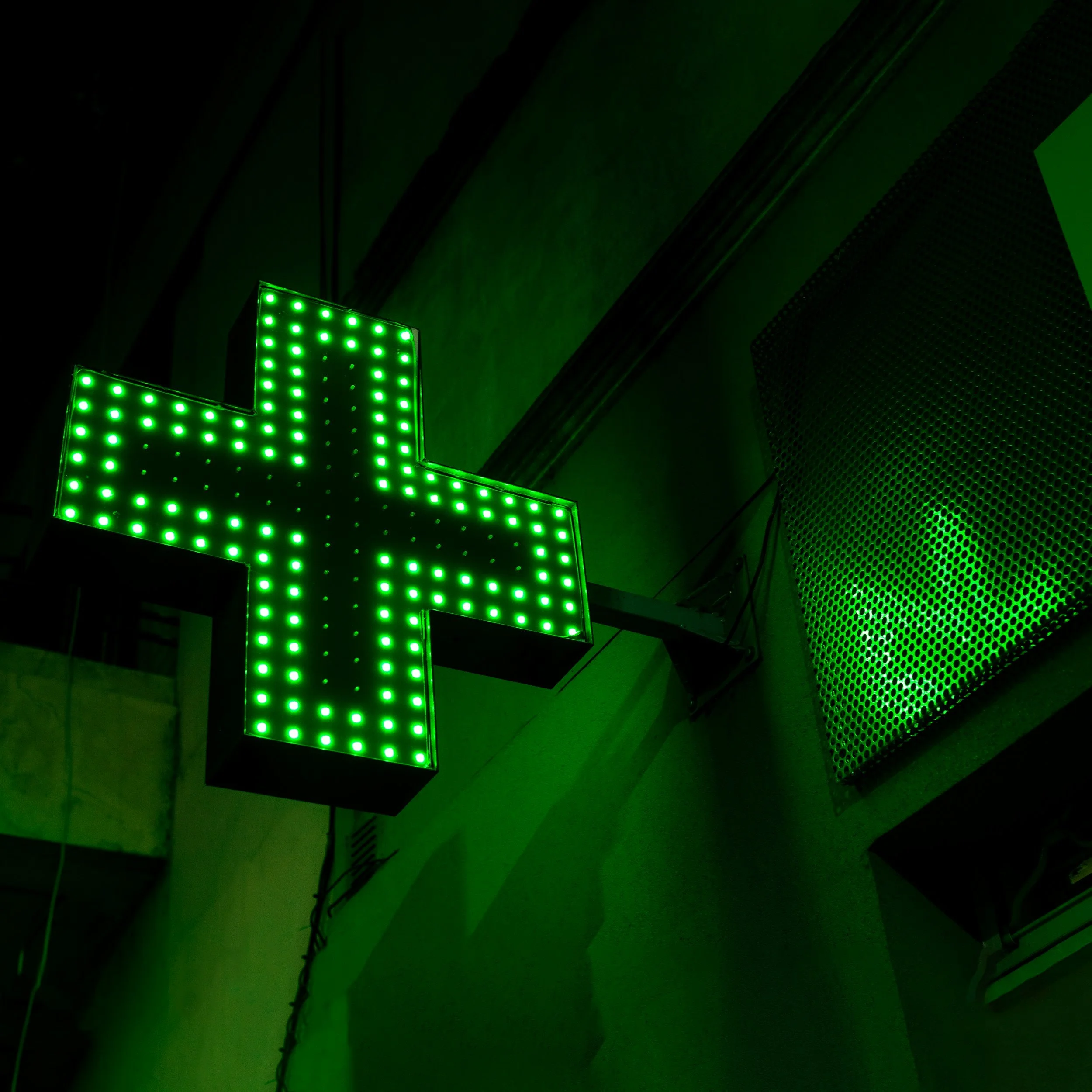 Green illuminated pharmacy cross sign on a dark building exterior.