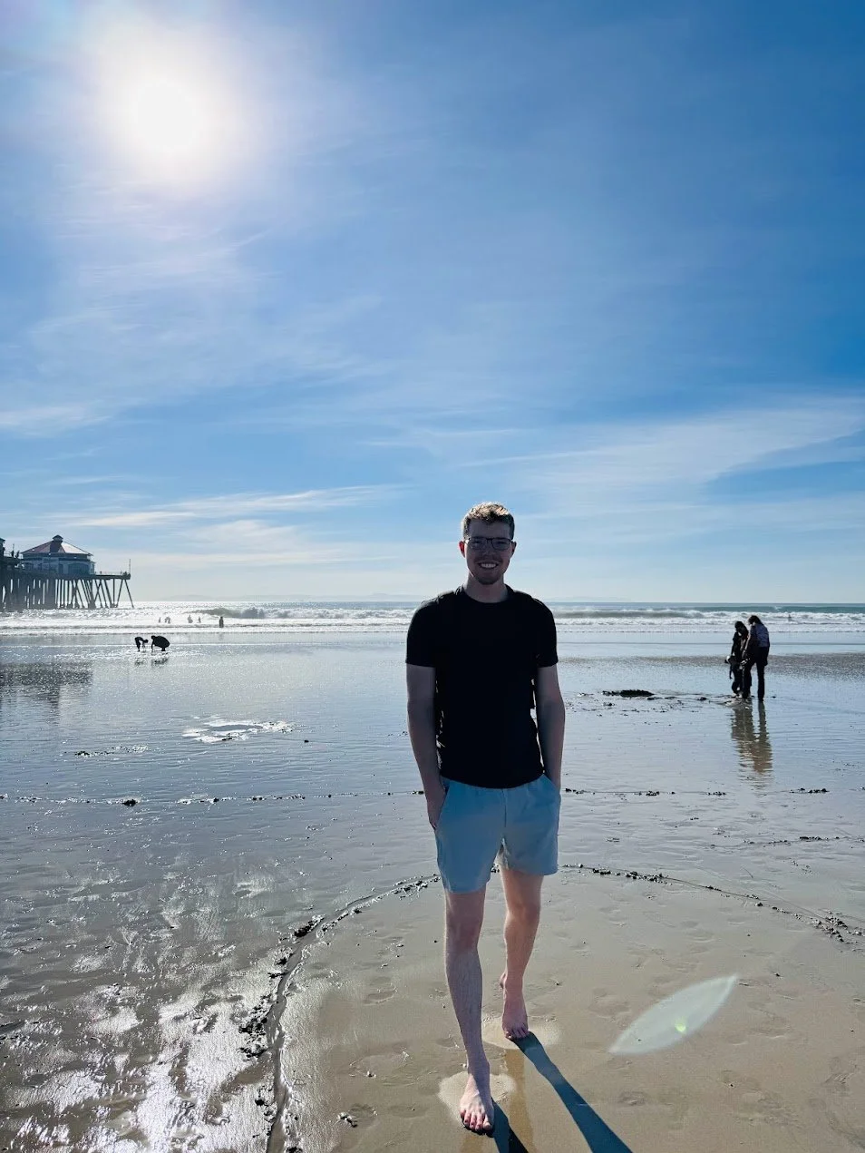 A smiling young man in black shirt and white shorts standing barefoot on a sandy beach, with a pier on the left and people in the background enjoying the beach under a clear blue sky.