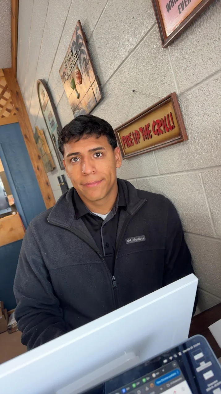 Young man with short dark hair, wearing a black Columbia jacket, sitting at a desk with a computer, in front of a beige cinder block wall decorated with framed signs and artwork, including a sign reading "Preheat the Grill!"