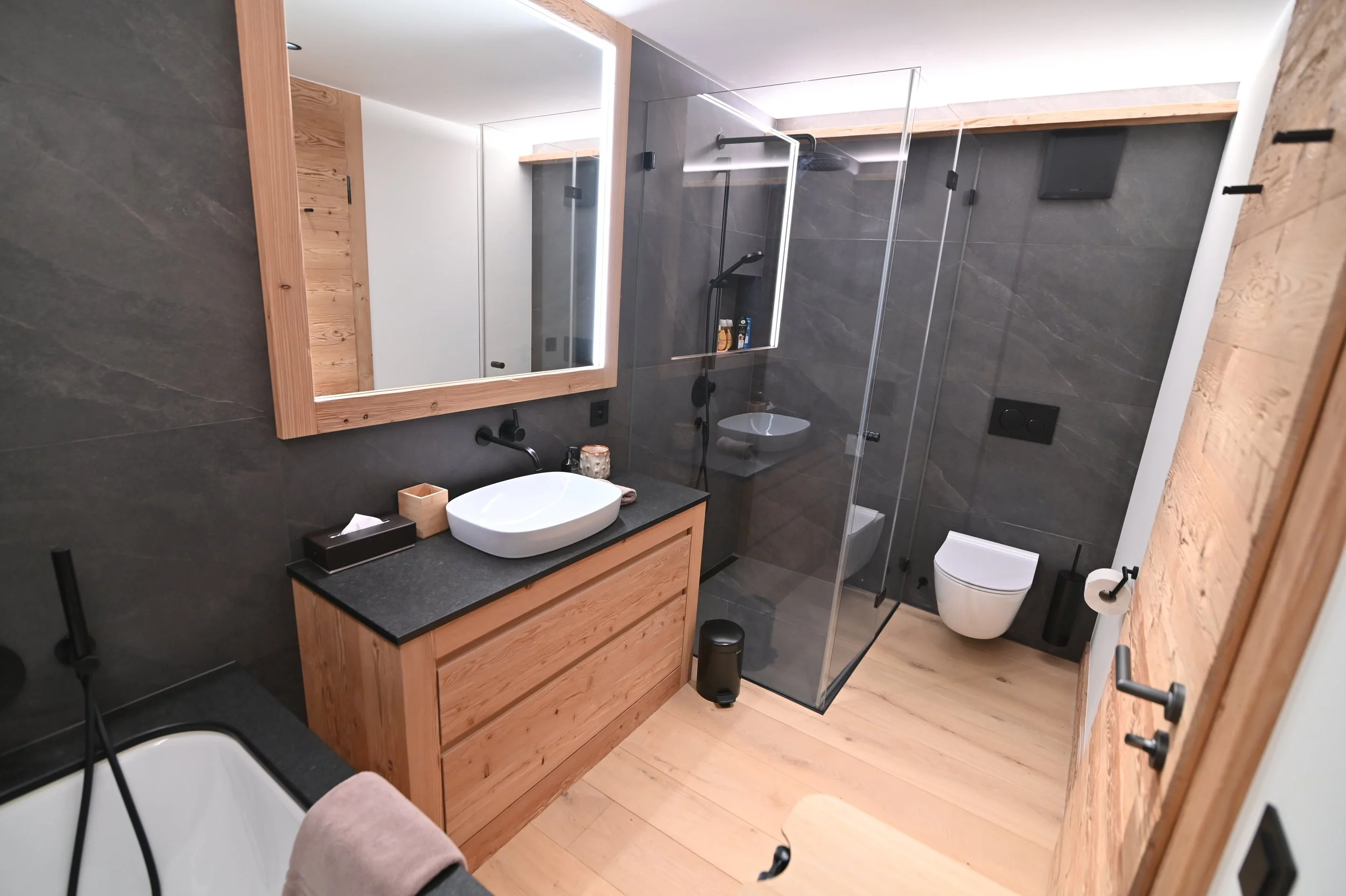 Ein modernes Badezimmer mit dunklen Wänden, Holzdetails und einer offenen Dusche mit Glastrennwand, einem Waschbecken auf Holzunterschrank, einem großen Spiegel, einem WC und Holzpanelen an der Wand.