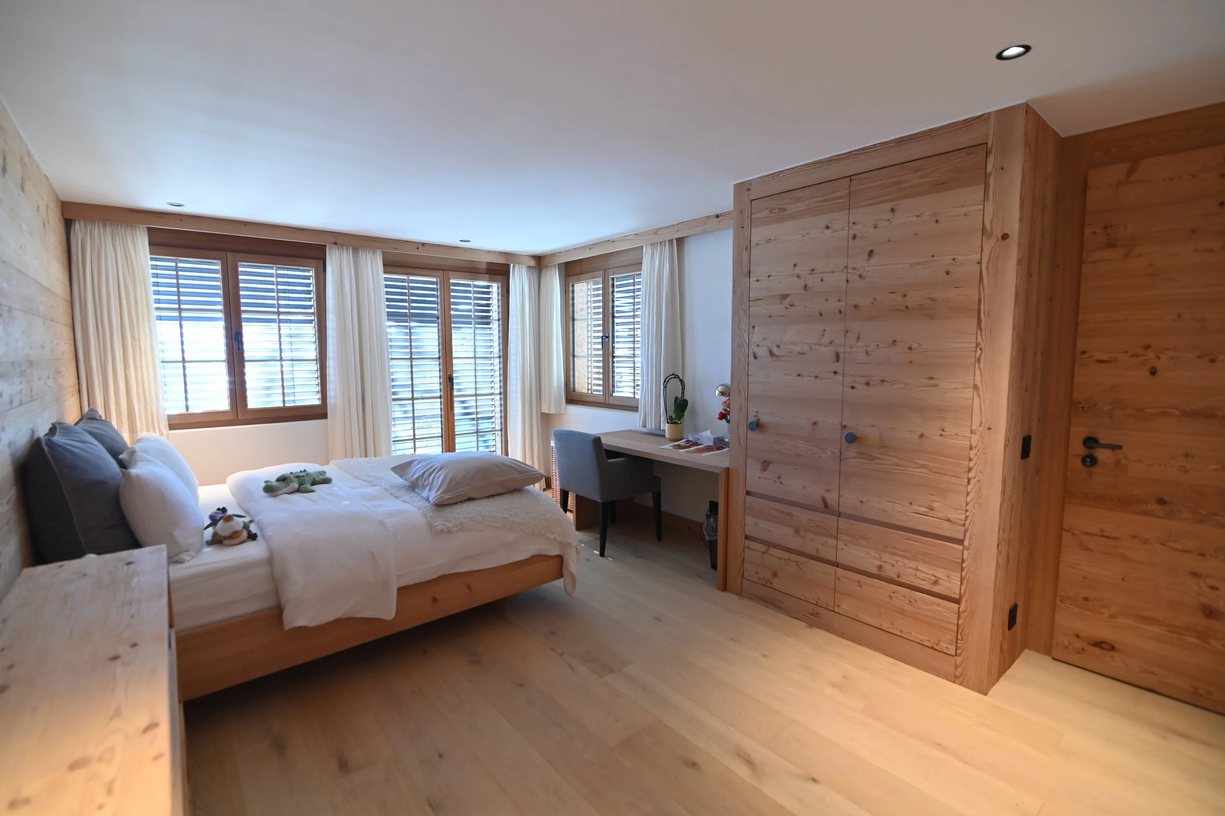Ein gemütliches Schlafzimmer mit einem Doppelbett, vielen Kissen und Plüschtieren. Es gibt große Fenster mit Vorhängen und eine Holzverkleidung an den Wänden und Möbeln.