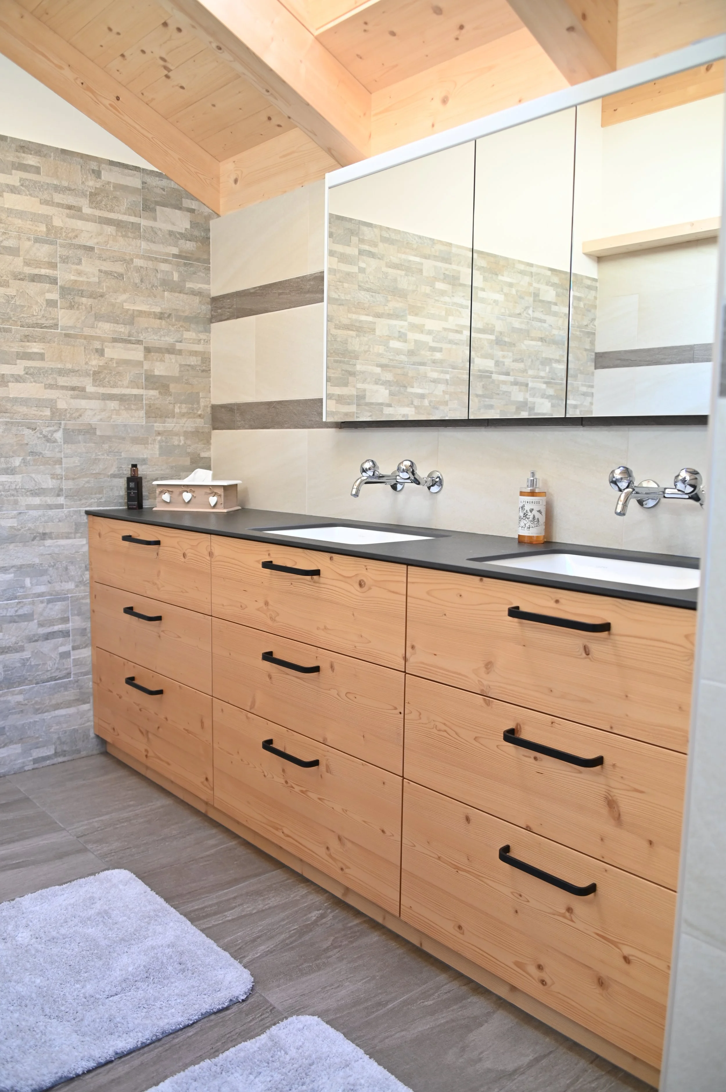 Ein modernes Badezimmer mit einem Holz-Unterschrank, einem großen Wandspiegel, Beckenwaschbecken, Wasserhähnen und einer Ablage mit Seife und Pflegeprodukten. Das Badezimmer hat eine Wand mit Steinfliesen und eine Decke aus Holz.