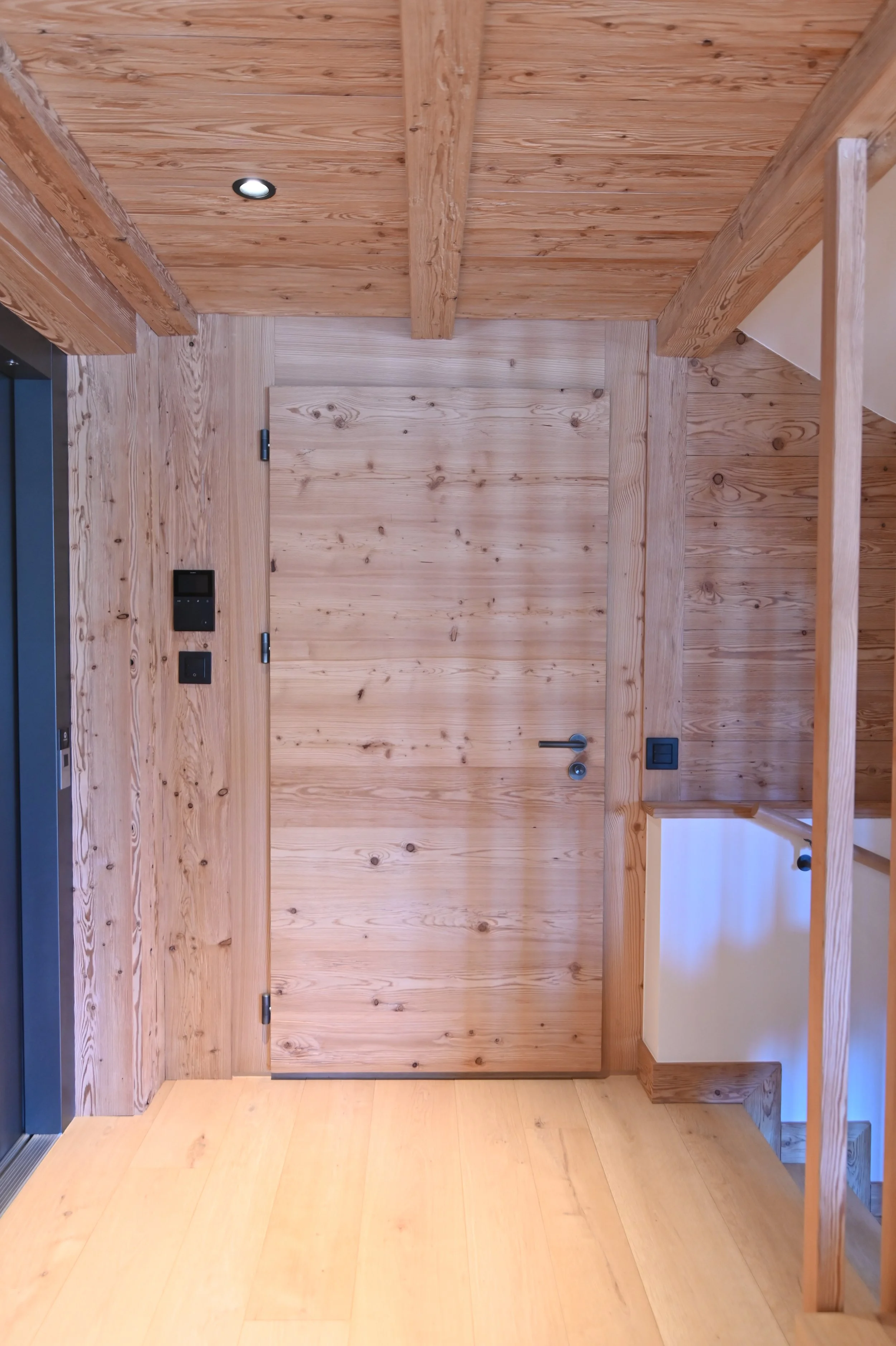 Holztür mit schwarzem Türgriff, sichtbare Holzvertäfelung an Wänden und Register, helles Holz, eingebauter Briefkasten und Lichtschalter.