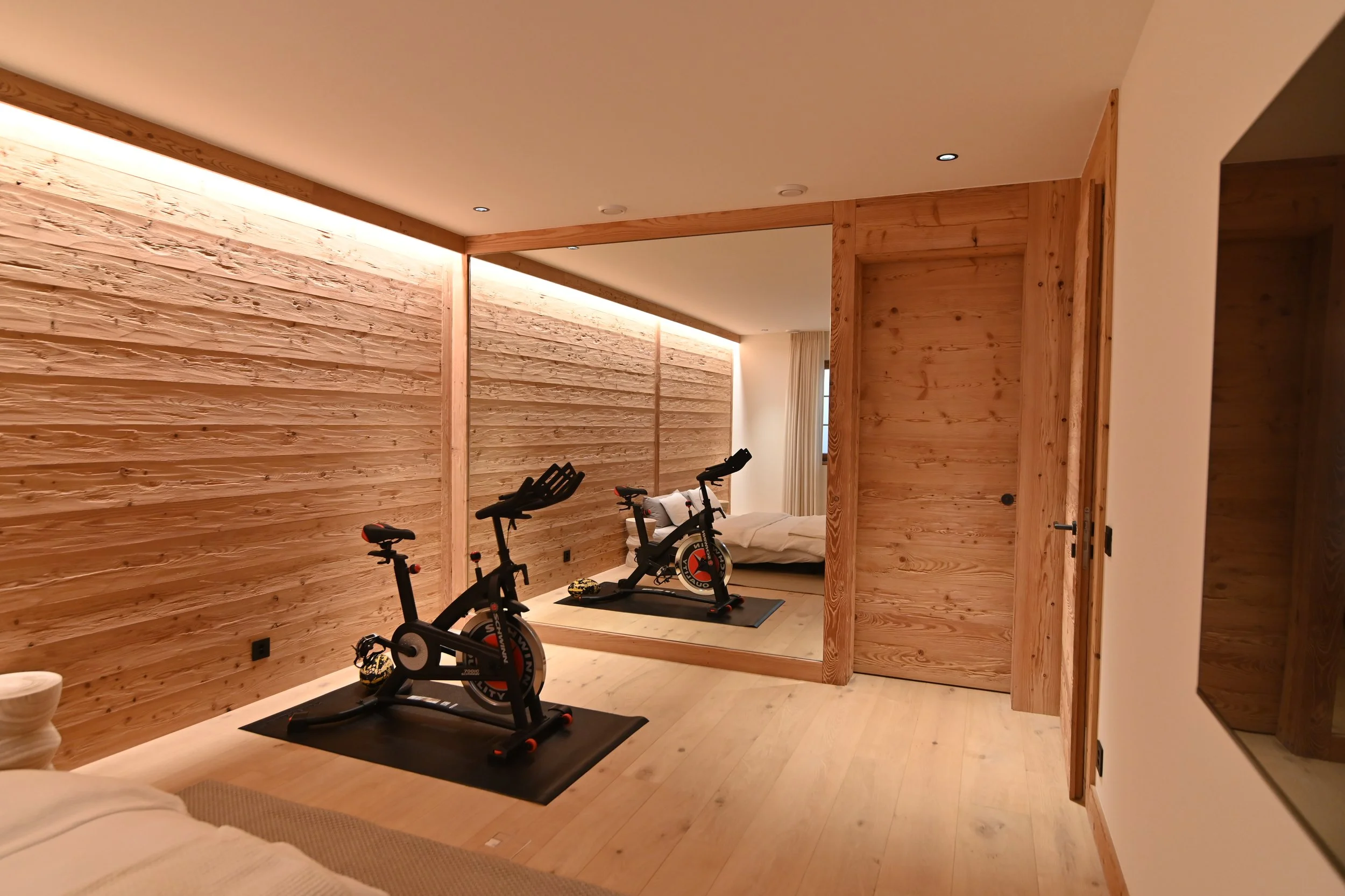 Ein Fitnessraum mit Holzvertäfelung, zwei Indoor-Fahrräder und ein Laufband auf einer Gymnastikmatte, beige Vorhänge vor einem Fenster, Deckenleuchten, Holztüren.