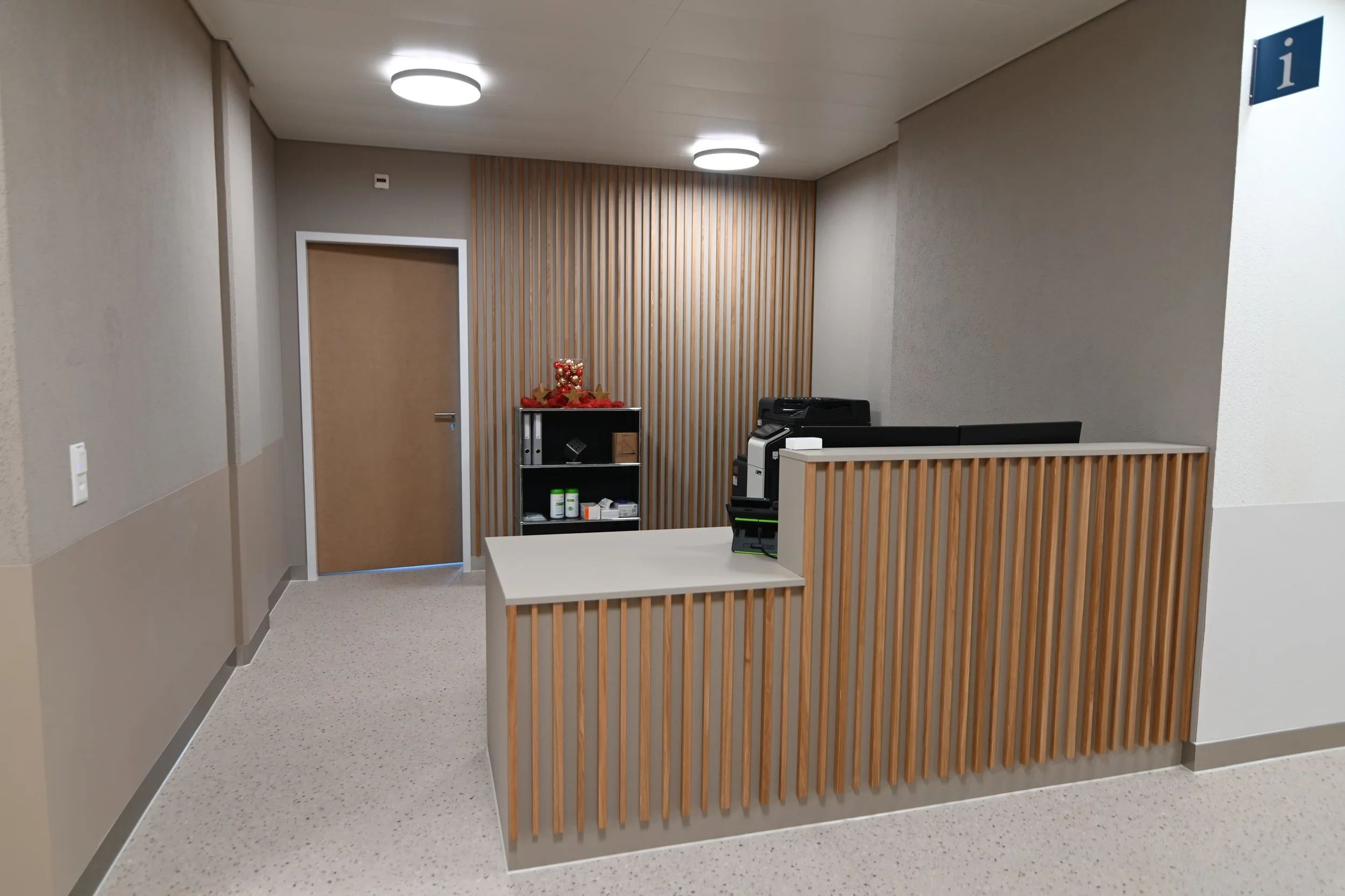 Innenansicht eines modernen Rathauses oder Büros mit Empfangstheke aus Holzlatten, einem beige Tür und einem Informationsschild ‚i‘ an der Wand.