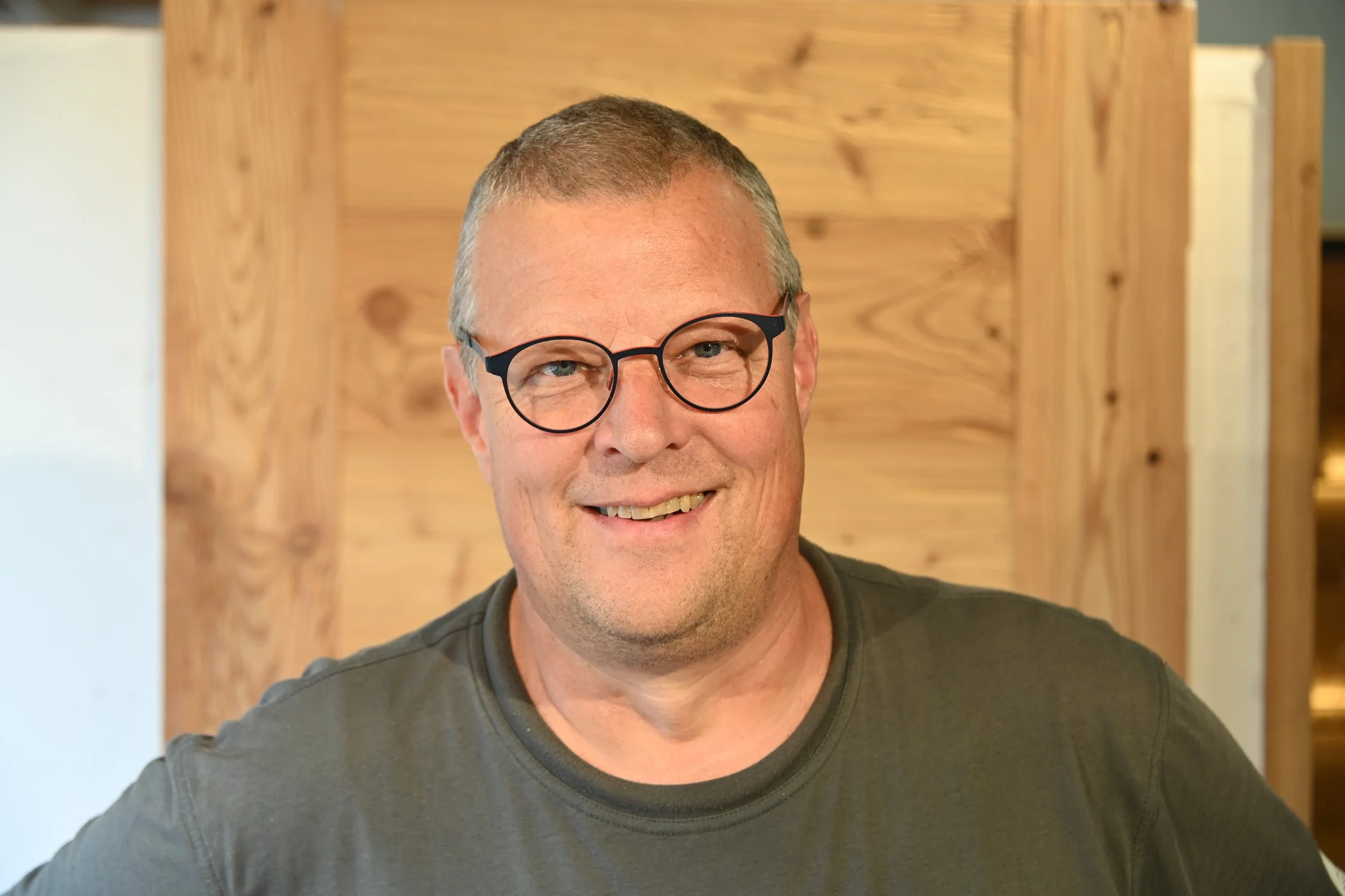 Nahaufnahme eines lachenden Mannes mit Brille und Kurzhaarfrisur vor einer Holzwand.