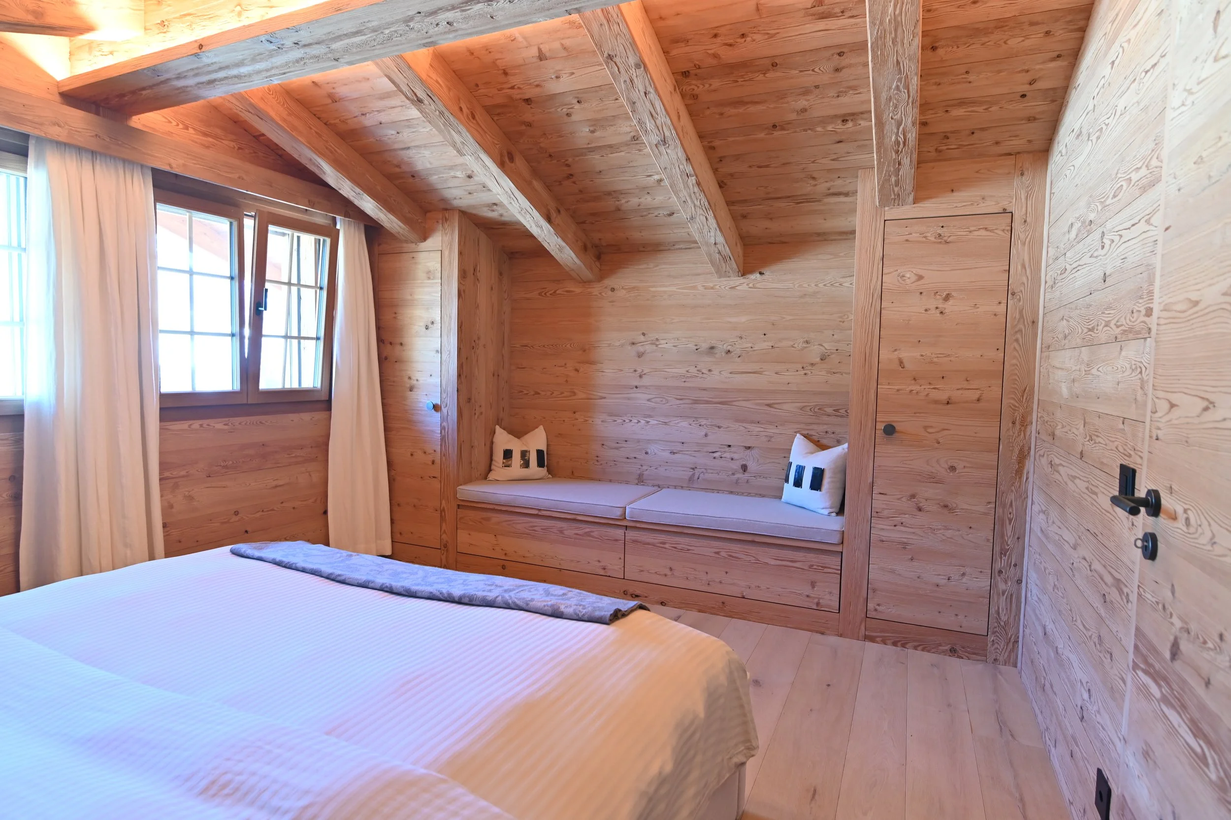 Innenraum eines wooden style Zimmers mit einem Bett, einem Sitzbereich mit Kissen und einer Fenster mit weißen Vorhängen.