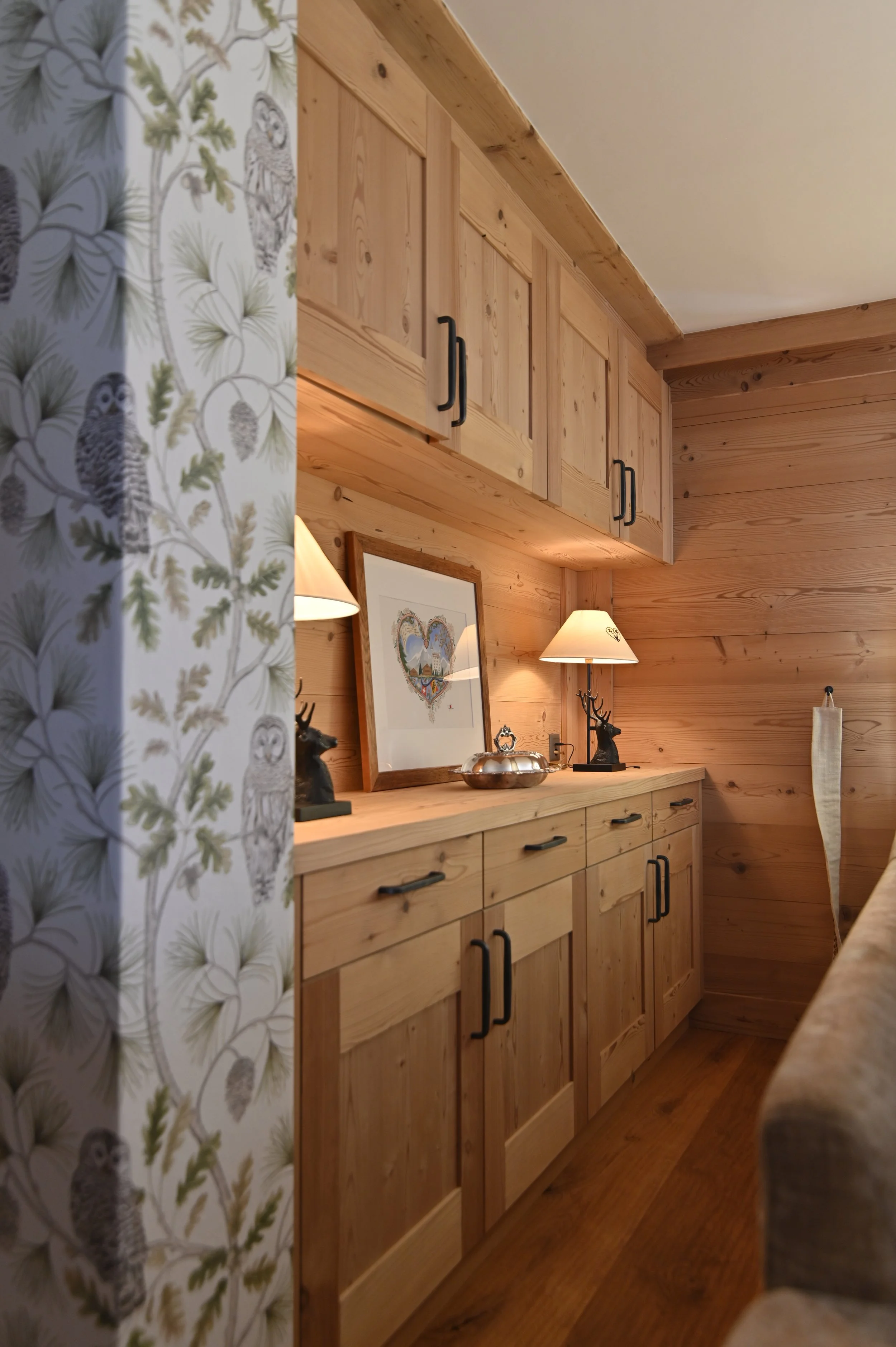Ein Holzschrank in einer rustikalen Holzwand mit schwarzen Griffen, Gegenstände auf einer Holzkommode darunter, eine Lampe und einen Rahmen, die Wand ist mit Holz verkleidet.