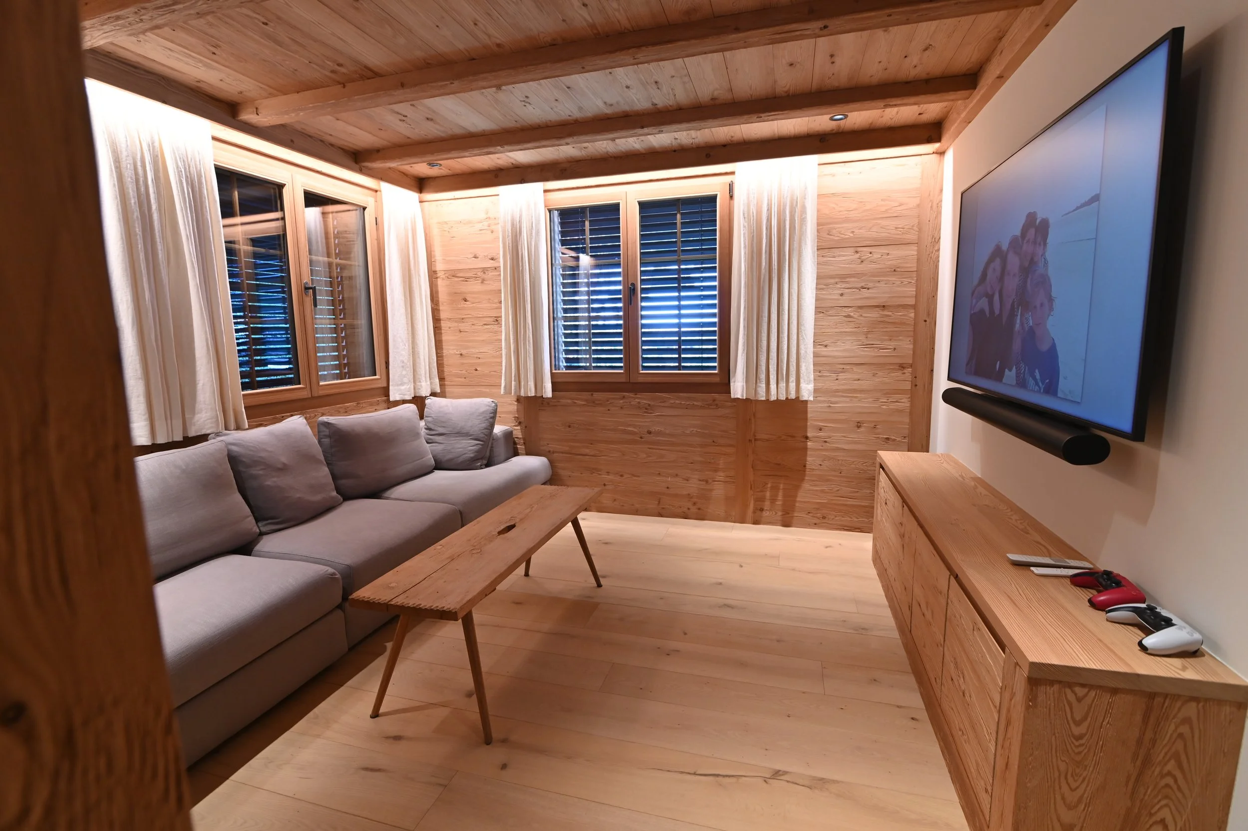 Wohnzimmer mit Holzdecke, Möbel, Fernseher, Fenster mit Vorhängen und Jalousien, Couch, Couchtisch, Spielecontroller.