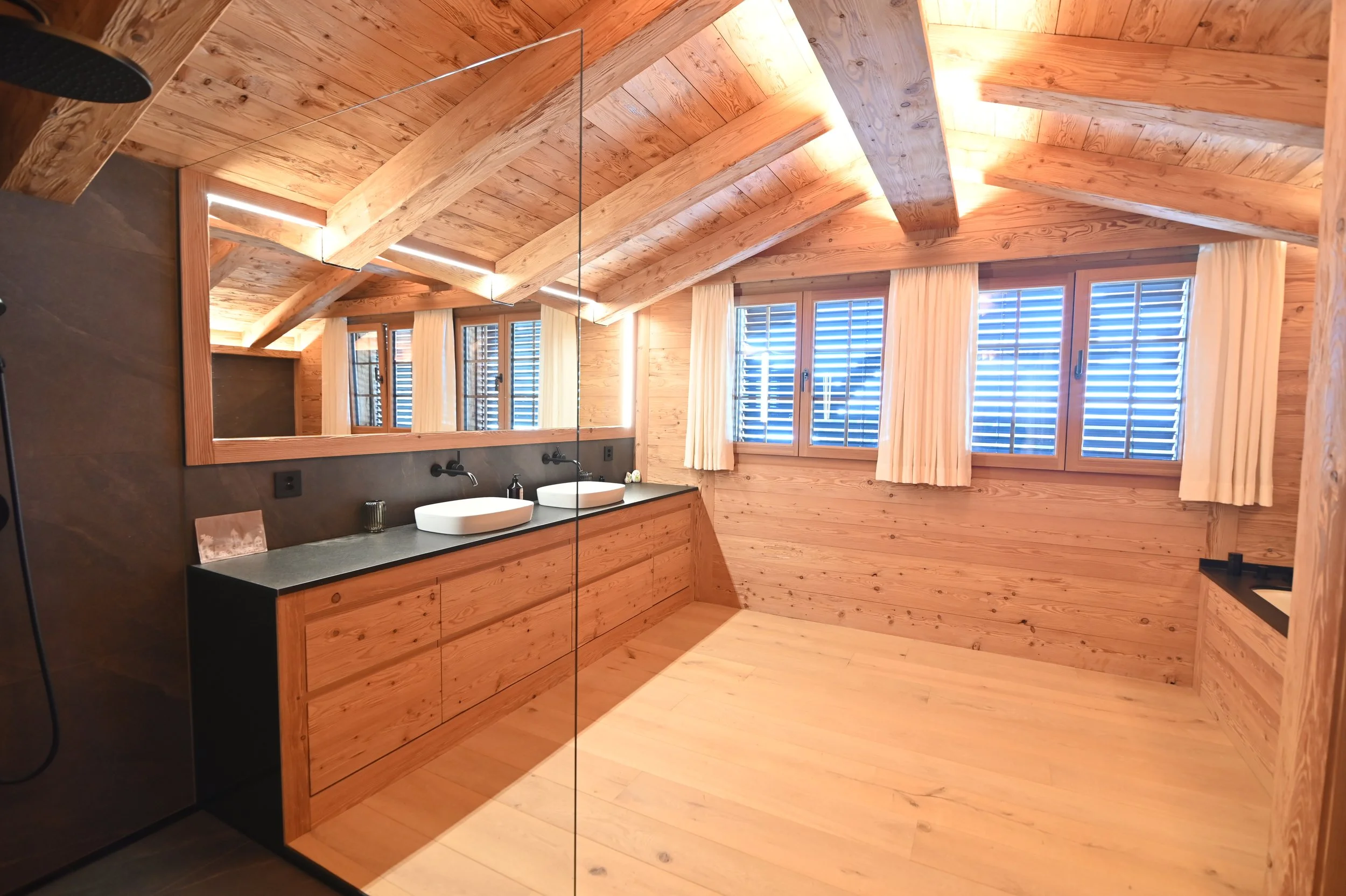 Holzverkleideter Badezimmerraum mit Doppelwaschbecken, großen Fenstern mit weißen Vorhängen und Spiegel an der Wand.