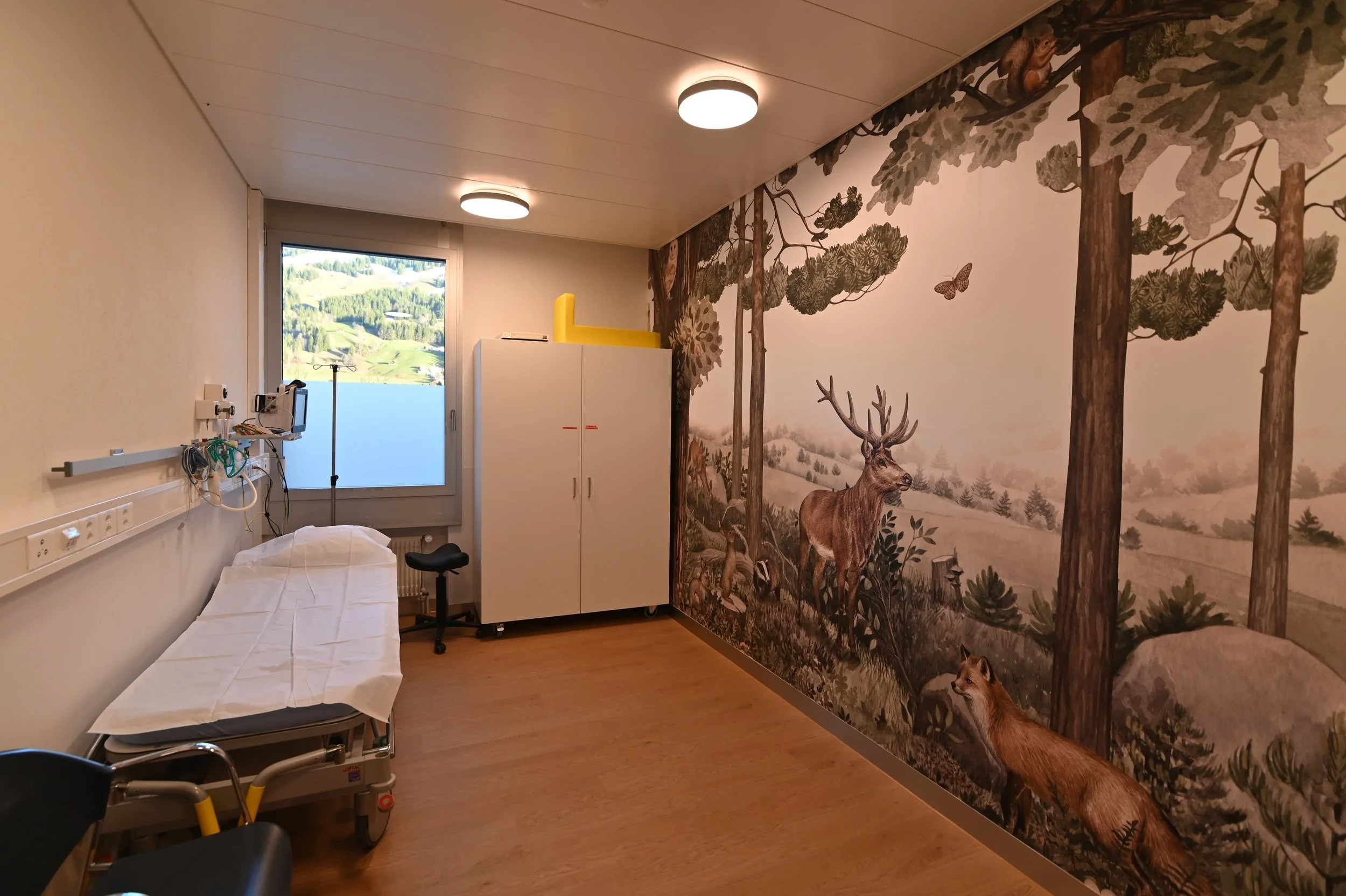 Ein Krankenhauszimmer mit einer Behandlungsliege, einem weißen Schrank, einer Fensteröffnung mit Blick auf eine grüne Landschaft und eine Wand mit Wald-Motiv mit Hirschen, Bäumen und Schmetterlingen.
