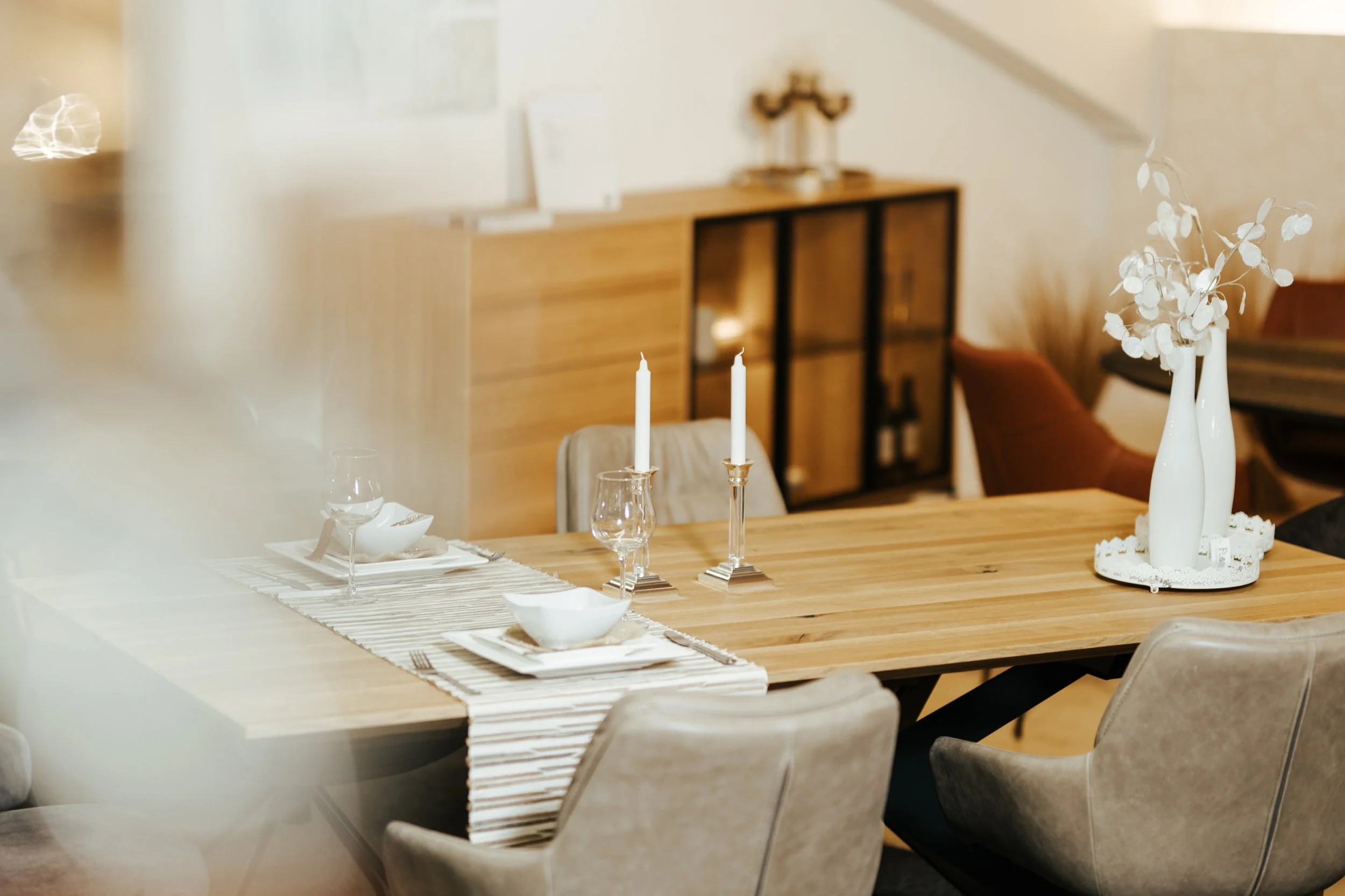 Ein Esstisch mit weißen Kerzen, einer weißen Vase mit weißen Blumen, weißen Tellern und Gläsern, in einem modern eingerichteten Raum mit Holzmobiliar und beige Stühlen.